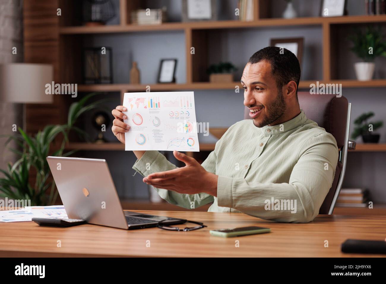 El joven hombre de negocios de carreras mixtas sonriente tiene una videollamada sentado en la oficina, mostrando el informe financiero con gráficos y tablas a colegas, socios y. Foto de stock