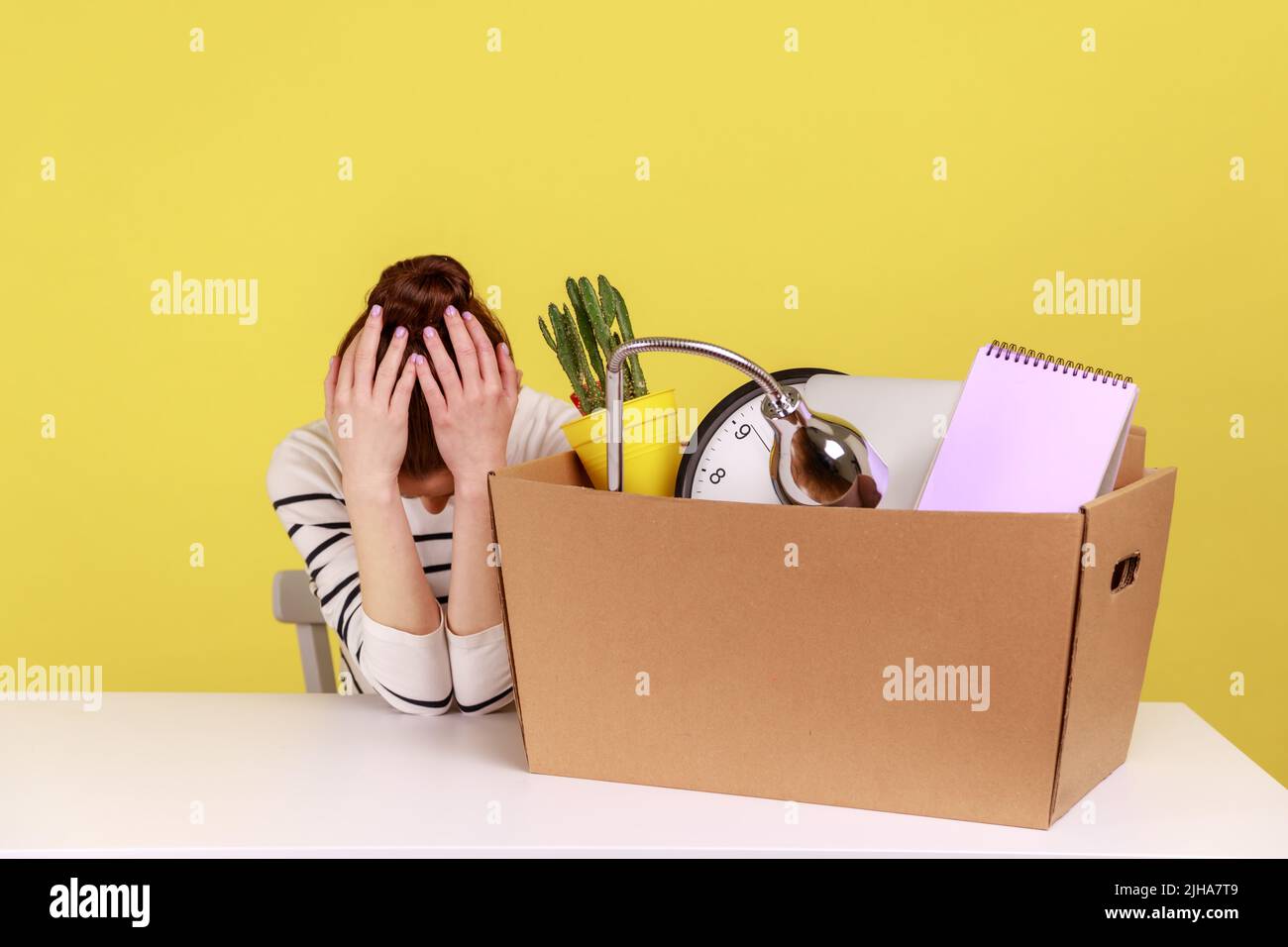 Triste, desesperada, joven trabajadora de oficina sentada en el lugar de trabajo con una caja de cartón con sus cosas, inclinó la cabeza desesperada. Estudio de interior grabado aislado sobre fondo amarillo. Foto de stock