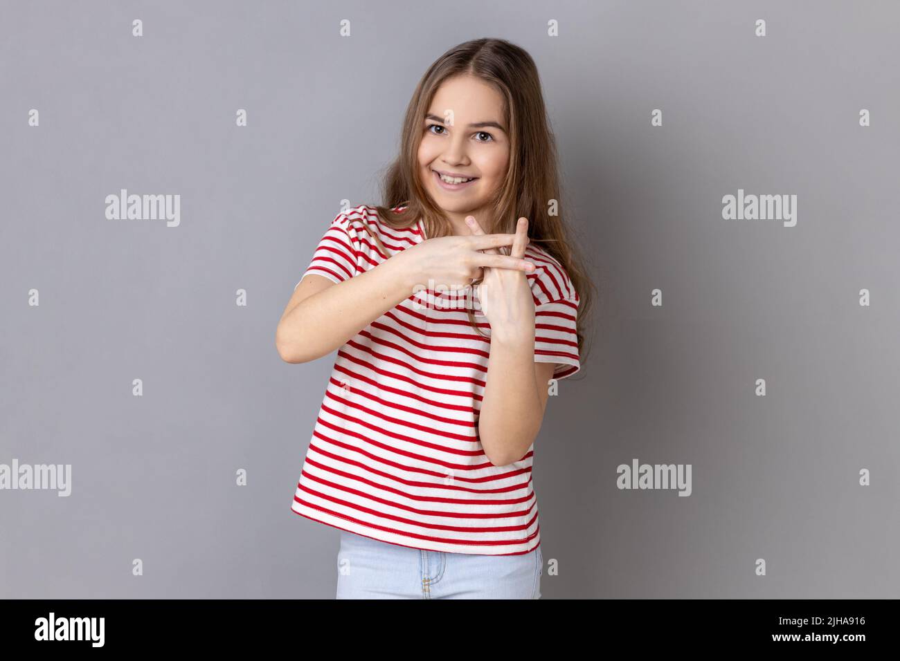 Tendencias de Internet. Retrato de niña que llevaba camiseta a rayas cruzando los dedos para hacer hashtag signo y mirar la cámara con una sonrisa tootea. Estudio de interior grabado aislado sobre fondo gris. Foto de stock