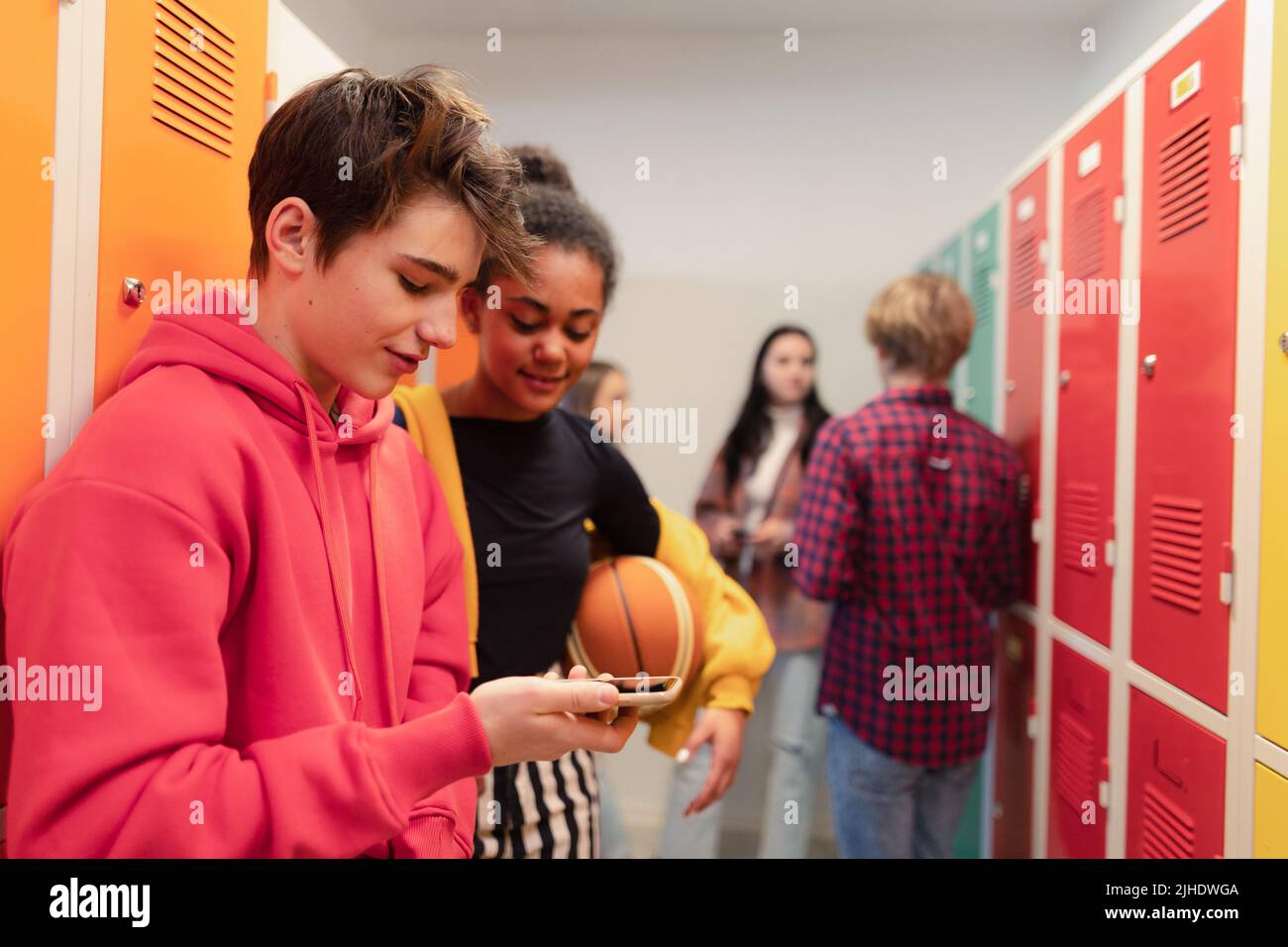 Jóvenes estudiantes de secundaria de pie cerca de un armario en el pasillo del campus hablando y utilizando el teléfono inteligente. Foto de stock