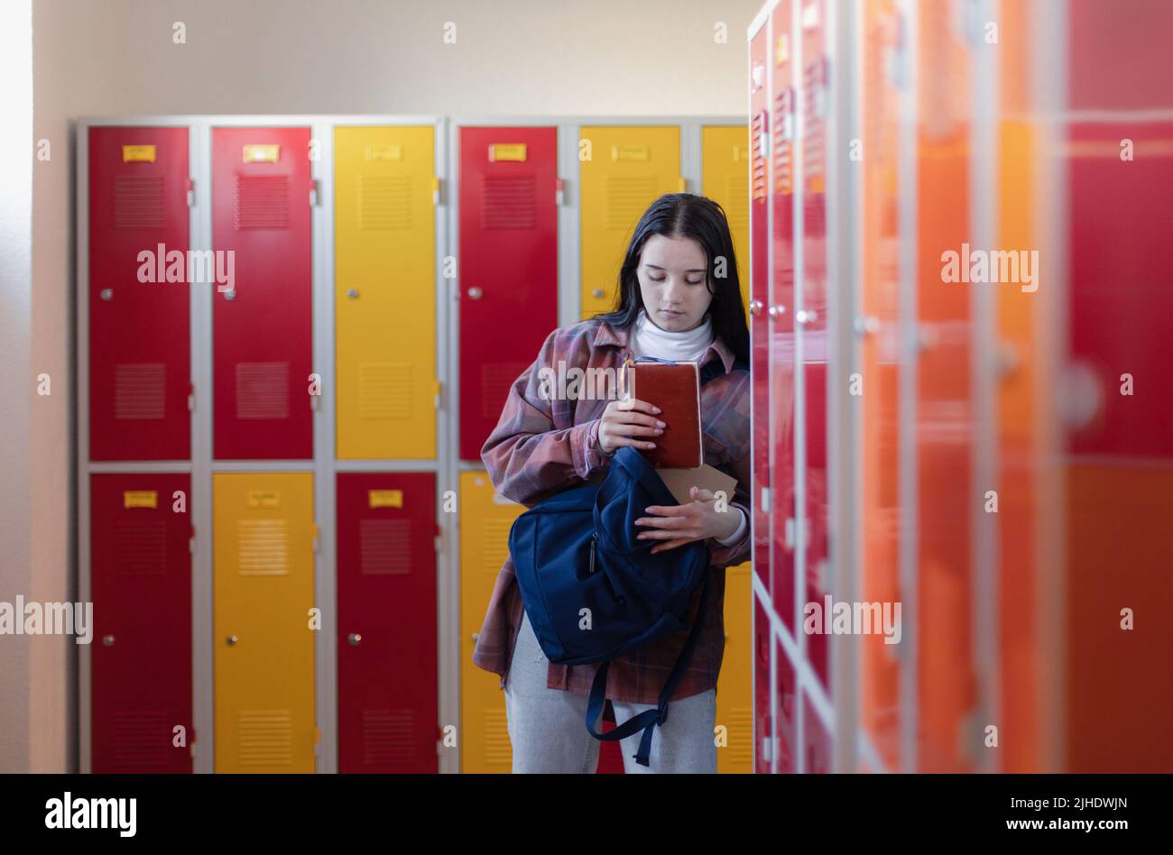 Estudiante adolescente triste parado en pasillo cerca de armarios coloridos y embalando libro a la mochila en pasillo del campus, concepto de vuelta a la escuela. Foto de stock