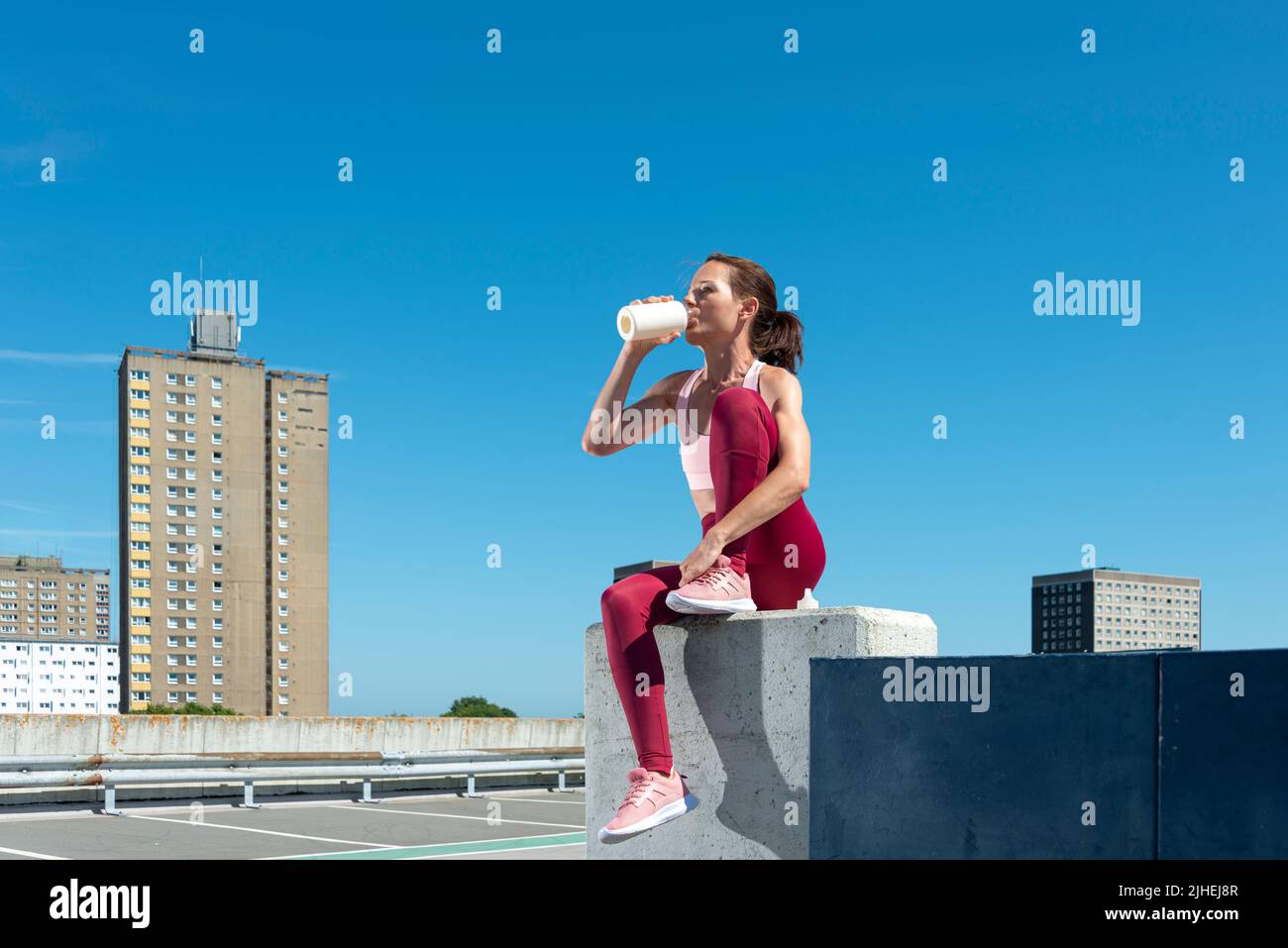 Mujer deportiva bebiendo agua de una botella, sentada descansando después del ejercicio, entorno urbano. Foto de stock