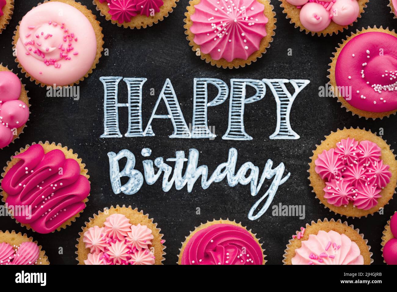 Vista superior de pastelitos rosados de cumpleaños dispuestos en una pizarra con un feliz saludo de cumpleaños escrito en tiza Foto de stock