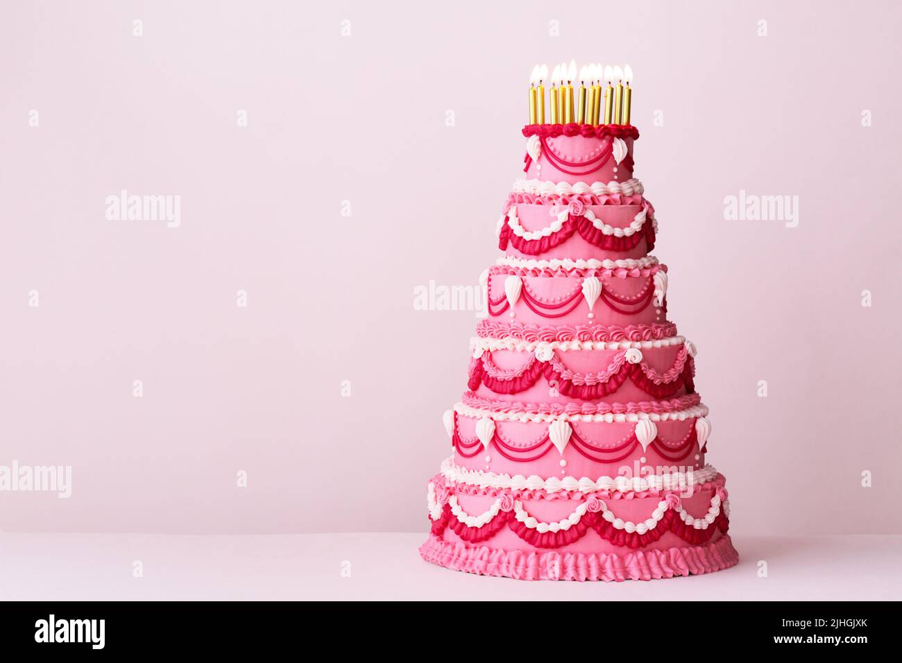 Extravagante pastel de cumpleaños de color rosa escalonado decorado con adornos de ribetes estilo crema vintage y velas de cumpleaños doradas Foto de stock