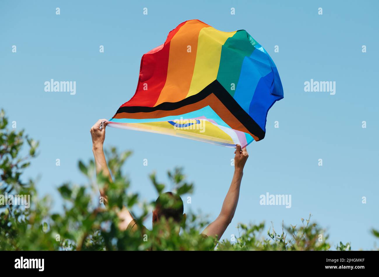 primer plano de un hombre joven, visto desde atrás, asomándose desde un arbusto, sosteniendo una bandera de orgullo de progreso inclusivo entre los sexos por encima de su cabeza Foto de stock