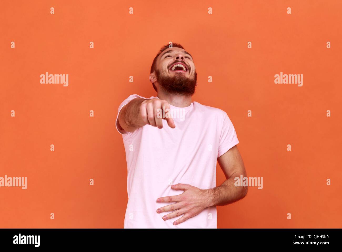 Retrato de hombre barbudo positivo gracioso riendo fuerte sosteniendo el vientre y señalando con el dedo en usted, burla, usando camiseta rosa. Estudio de interior grabado aislado sobre fondo naranja. Foto de stock