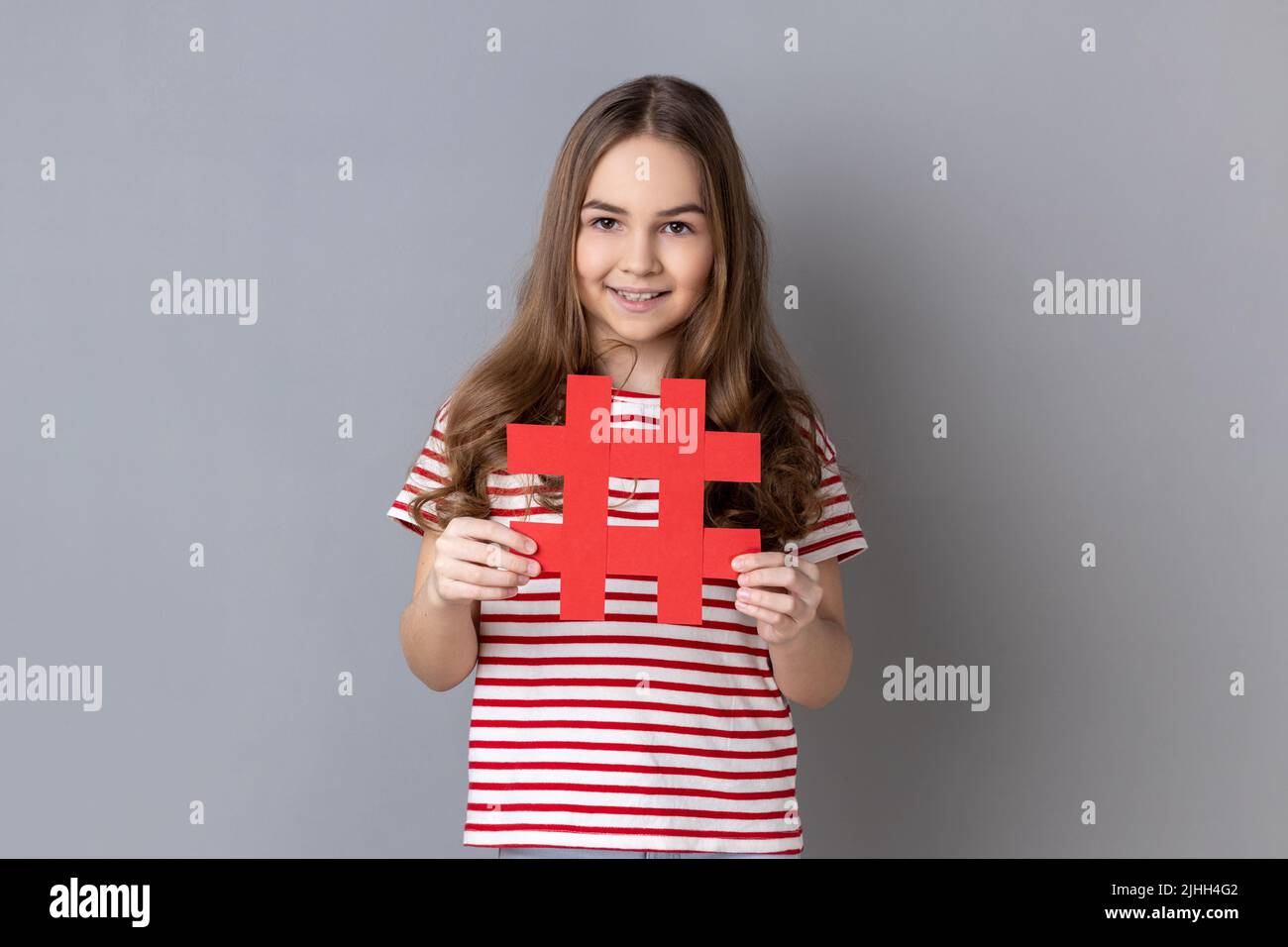 Retrato de niña feliz con camiseta a rayas que sostiene hashtag símbolo, la promoción de tema viral en la red social, marcado de tendencias de blog. Estudio de interior grabado aislado sobre fondo gris. Foto de stock