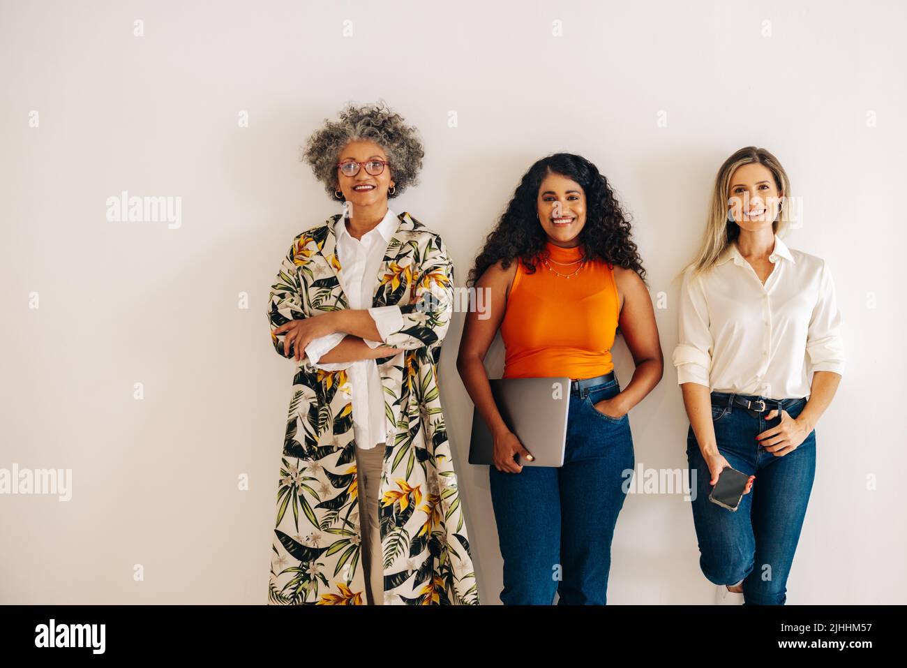 Tres mujeres de negocios diversas sonriendo a la cámara mientras se encontraban de pie contra una pared de oficina. Grupo de empresarias multiculturales que trabajan juntas en una A. Foto de stock
