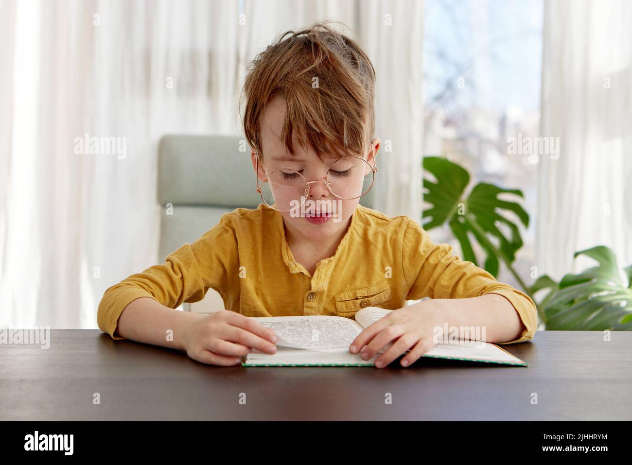 Niño sentado en la silla leyendo un libro, usando gafas Foto de stock