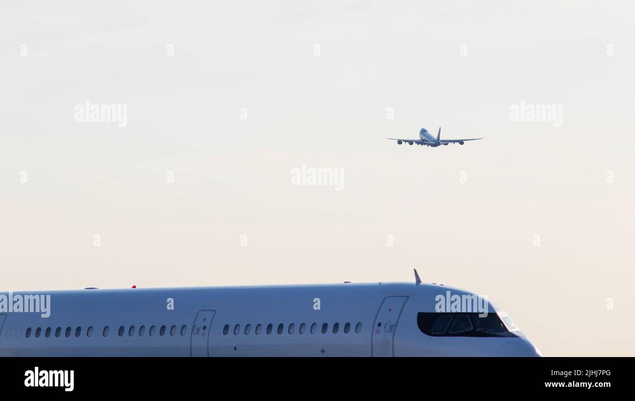 Un avión de pasajeros se ve despegando en el fondo, mientras que otro avión se ve pasar, de cerca en una mañana despejada. Foto de stock