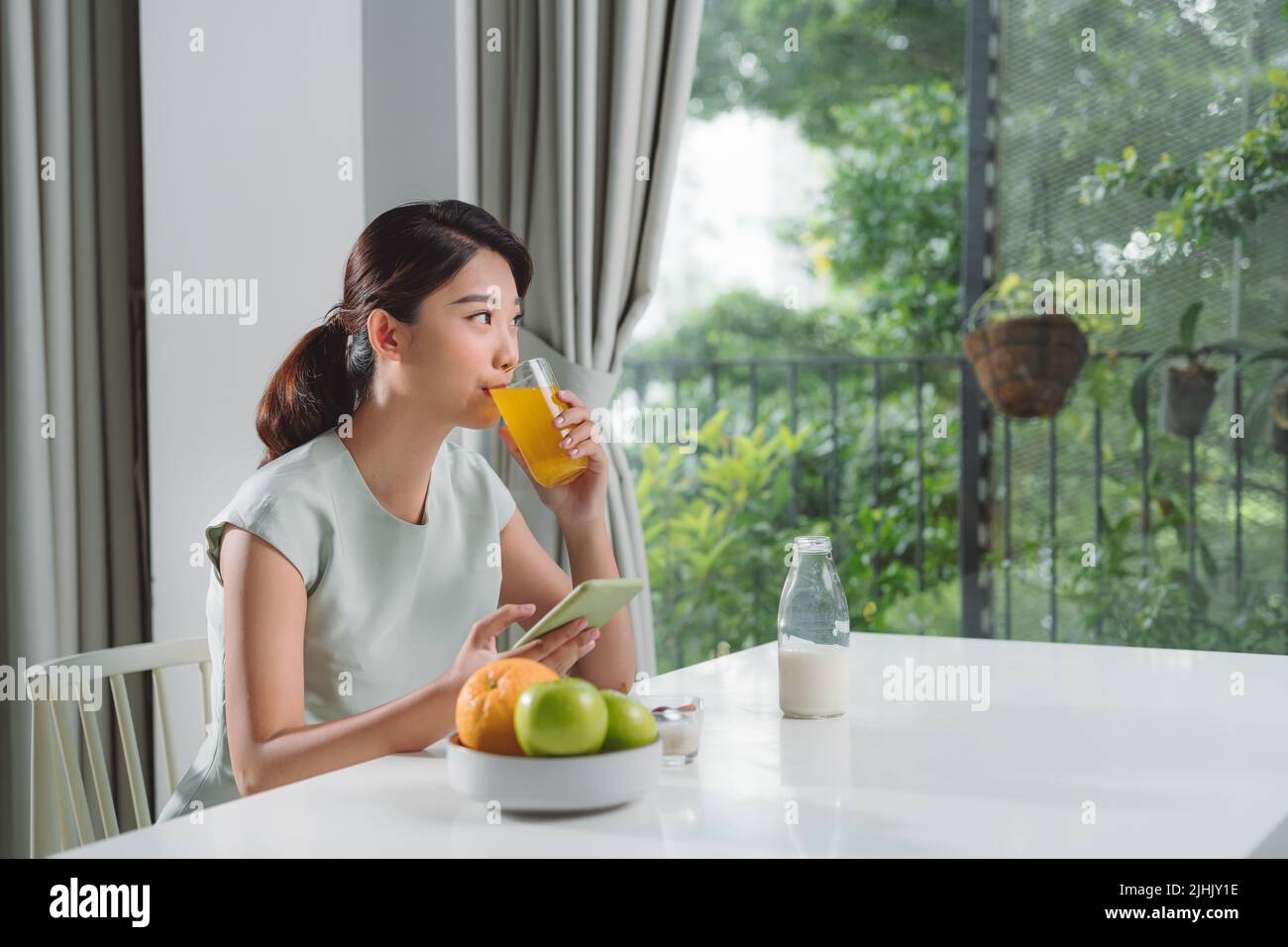 mujer tomando jugo de naranja mientras usa el teléfono móvil en casa Foto de stock