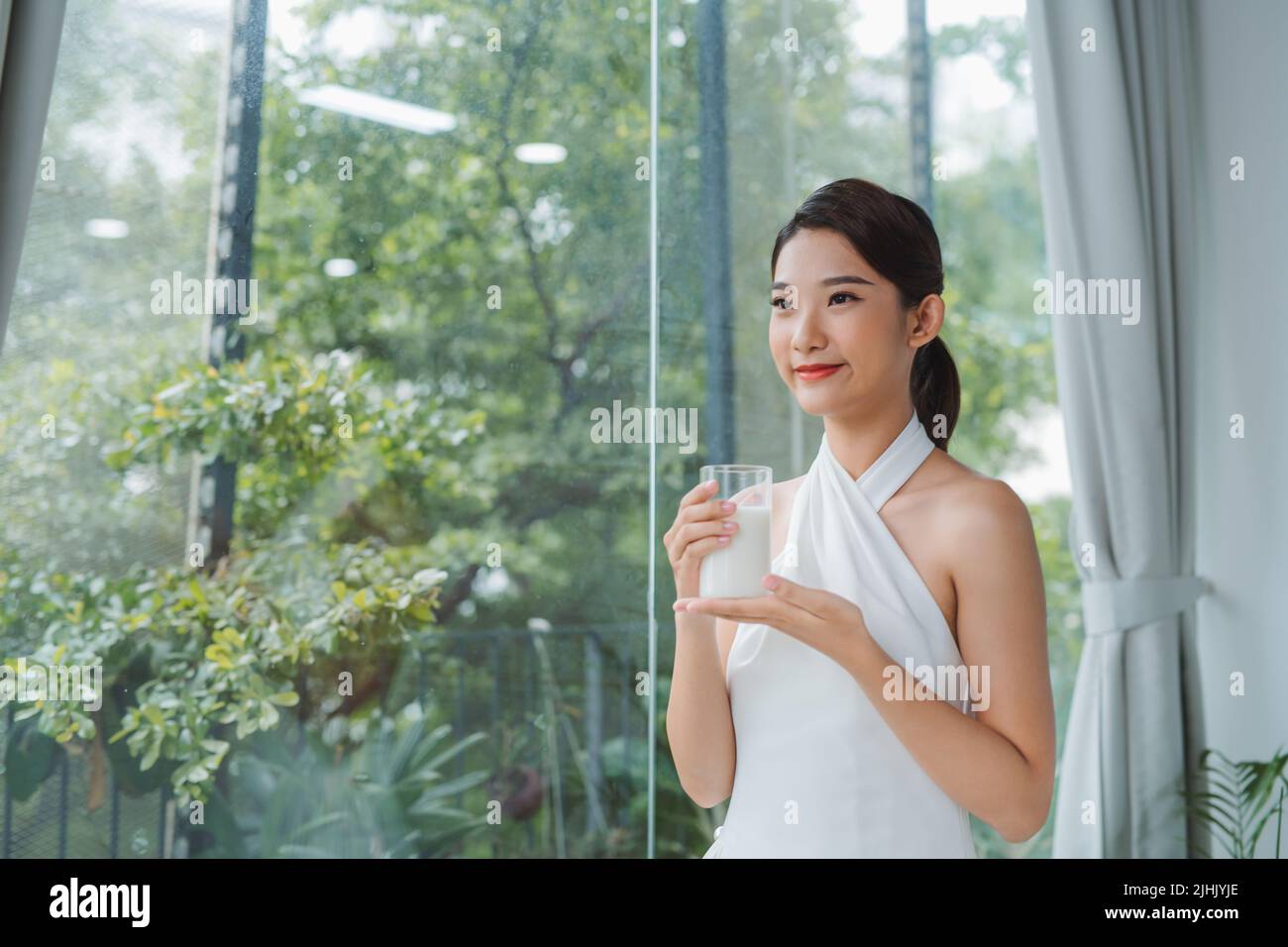 Mujer asiática bebiendo un vaso de leche mirando por la ventana Foto de stock