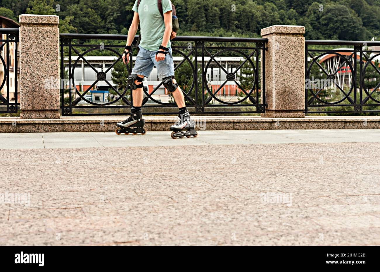 Hombre joven con ropa azul en equipo de protección montado sobre patines a lo largo del terraplén contra el fondo del bosque y las montañas en summe Foto de stock