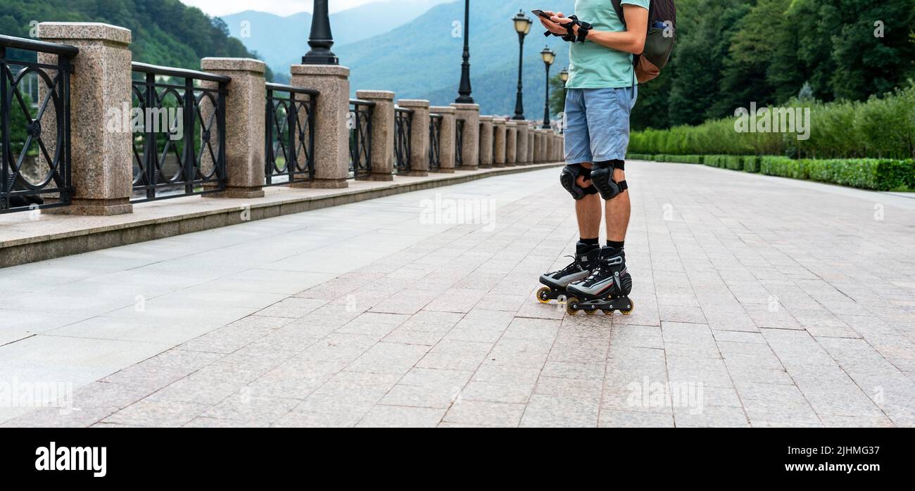 hombre joven en ropa azul en equipo de protección montado sobre patines con smartphone en verano actividades al aire libre modernas tecnologías Foto de stock
