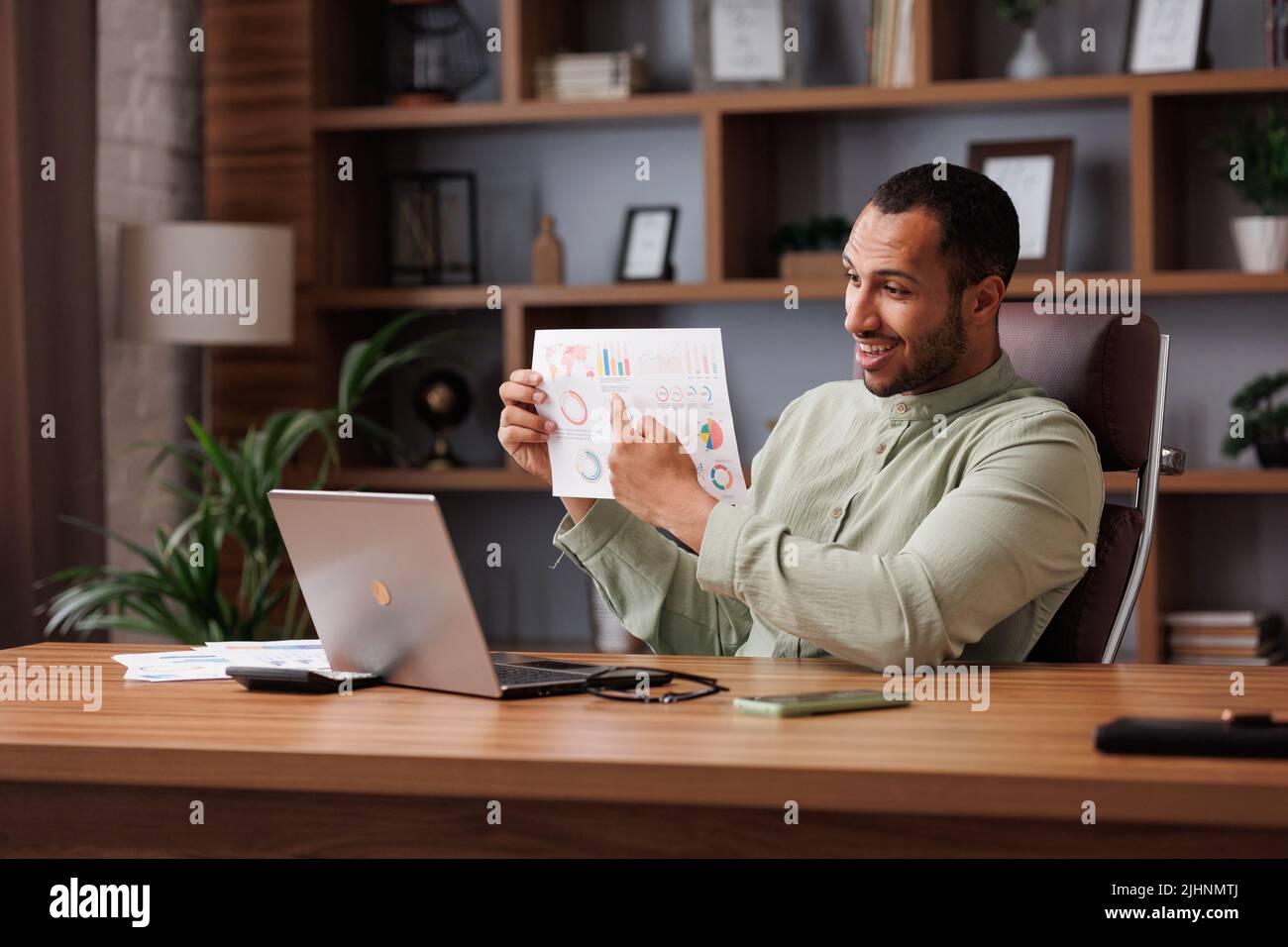 El joven hombre de negocios de carreras mixtas sonriente tiene una videollamada sentado en la oficina, mostrando el informe financiero con gráficos y tablas a colegas, socios y. Foto de stock