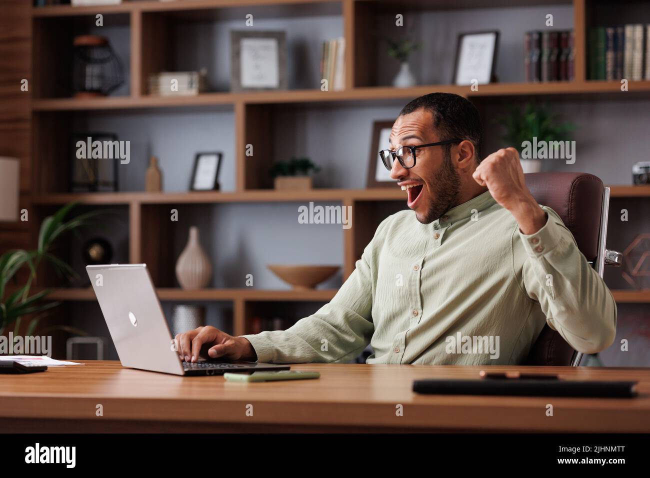 Feliz hombre de negocios afroamericano sorprendido por las buenas noticias y haciendo un gesto de sí mientras mira el ordenador portátil sentado en la oficina. El gerente disfruta del éxito Foto de stock
