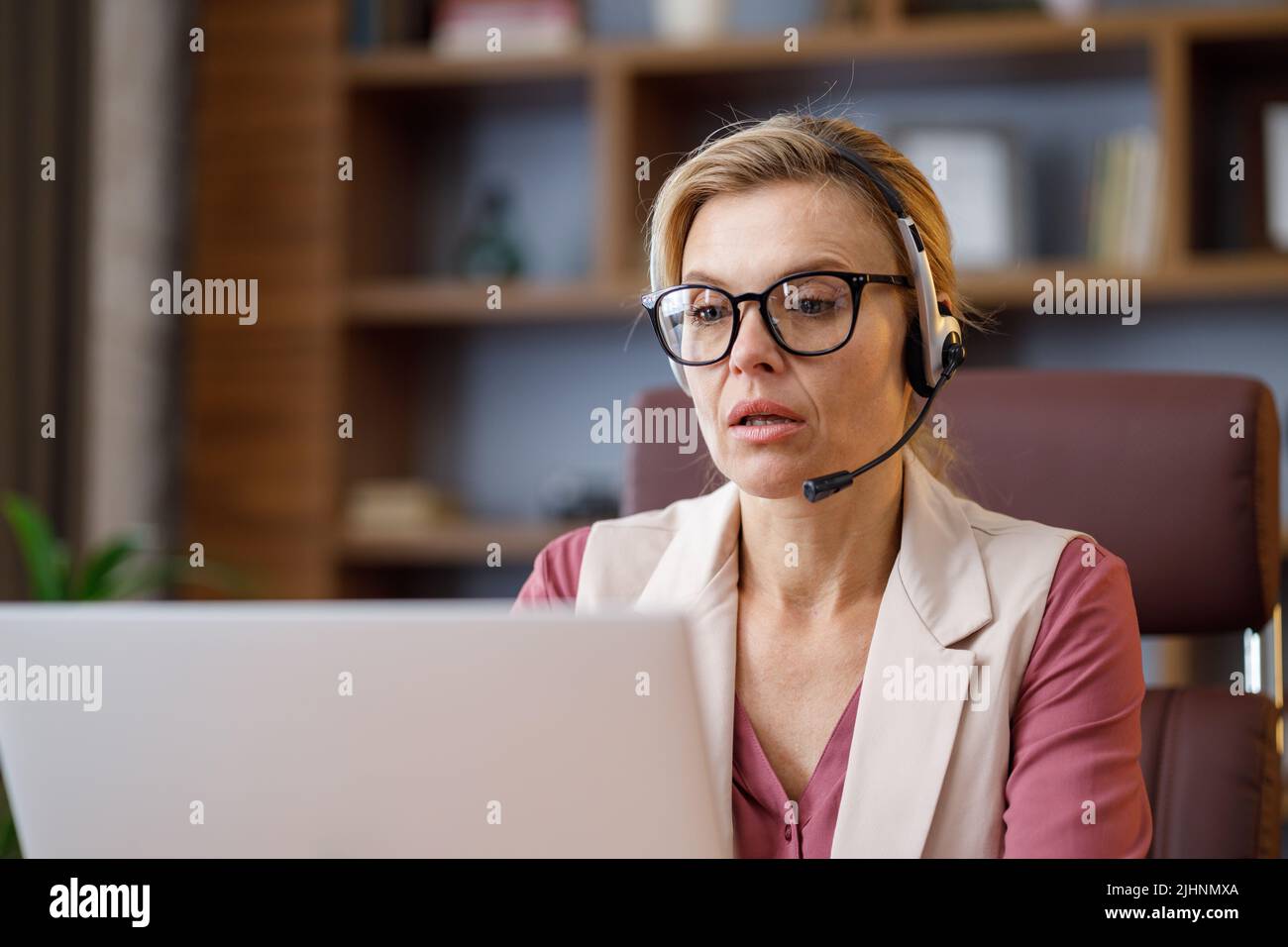 Mujer de negocios sonriente con auriculares hablando con el portátil cámara web sentada en la oficina. Grabación de vlog, videoconferencia en línea, videollamada, en línea Foto de stock