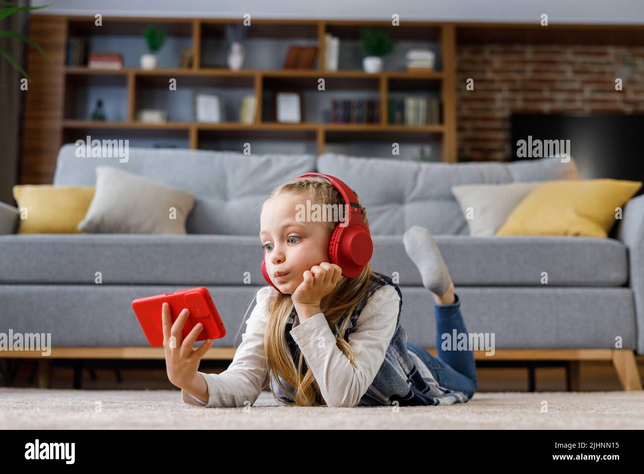 Niña sorprendida con auriculares rojos tumbada en la alfombra de casa. Chica linda sosteniendo smartphone, viendo dibujos animados, mensajes de texto, navegando Foto de stock