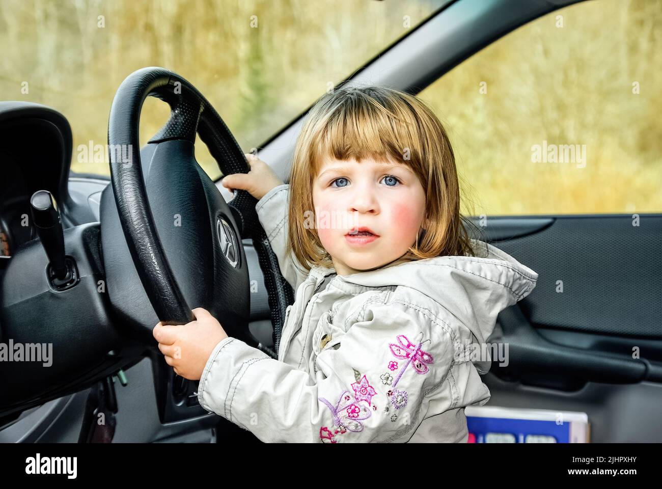 Una niña pequeña, una niña está en el volante de un coche jugando a la conducción y mira a la cámara, atrapado, pero también seguro de sí mismo. Foto de stock