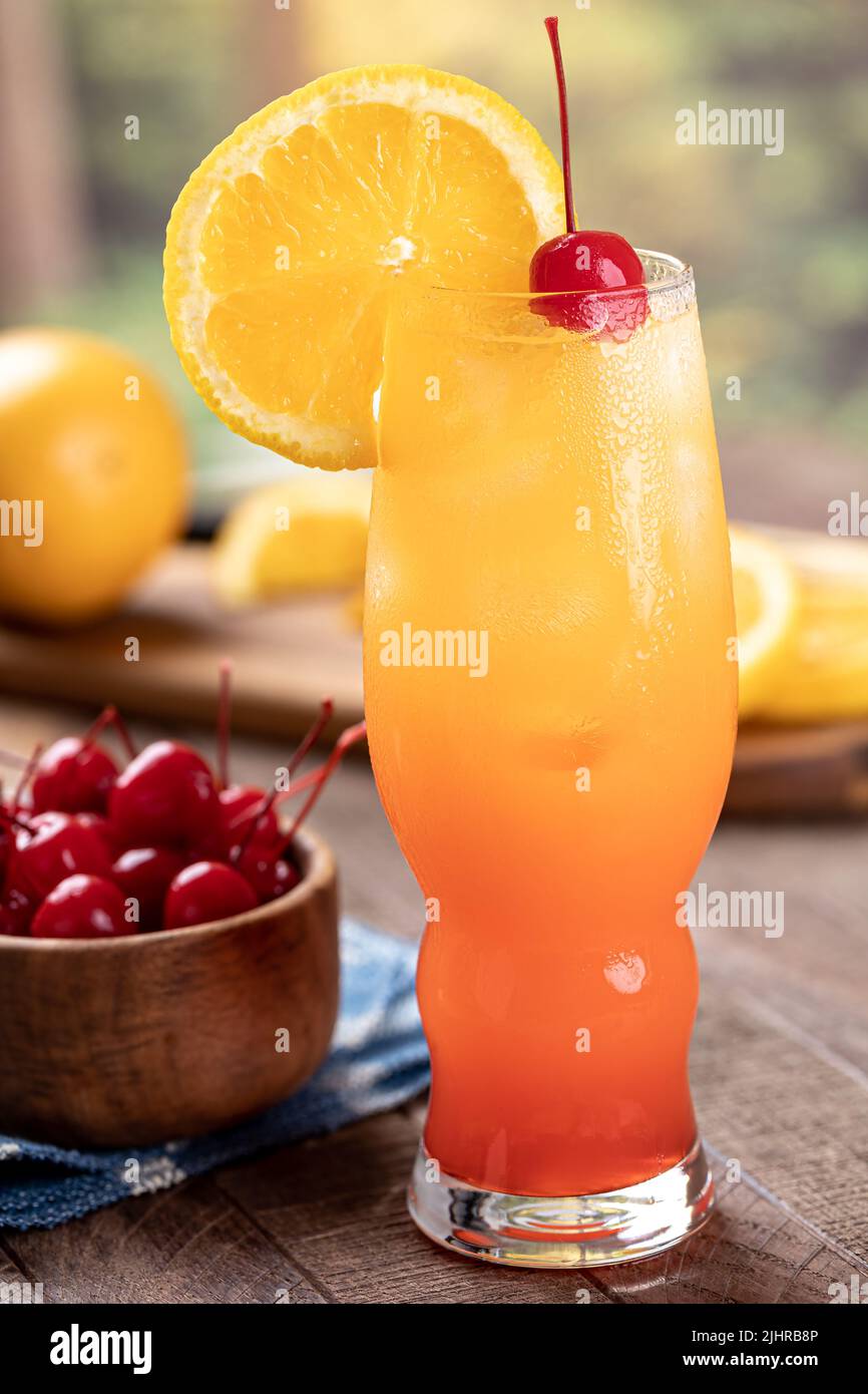 Cóctel Tequila Sunrise con rodaja de naranja y cereza al aire libre sobre una mesa rústica de madera Foto de stock