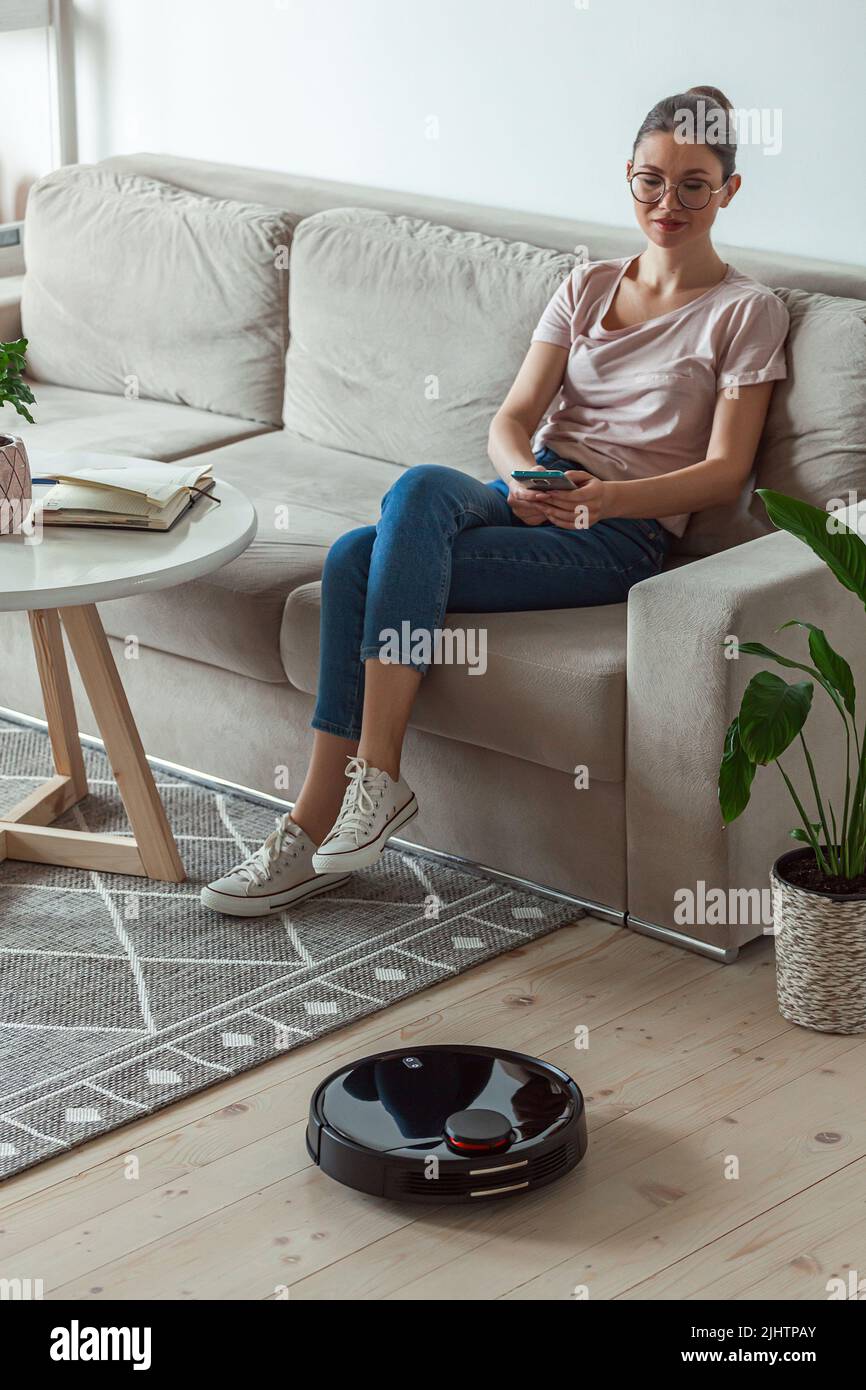 Aspiradora robot limpieza moqueta, mujer mando a distancia teléfono móvil y  disfrutar de descanso, sentado en el sofá en casa Fotografía de stock -  Alamy