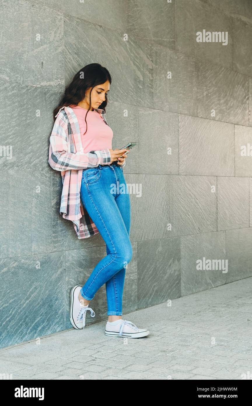 Una joven adolescente muy de moda charlando con su teléfono móvil en las redes sociales mientras se apoya contra una pared gris en retroceso con copyspace Foto de stock