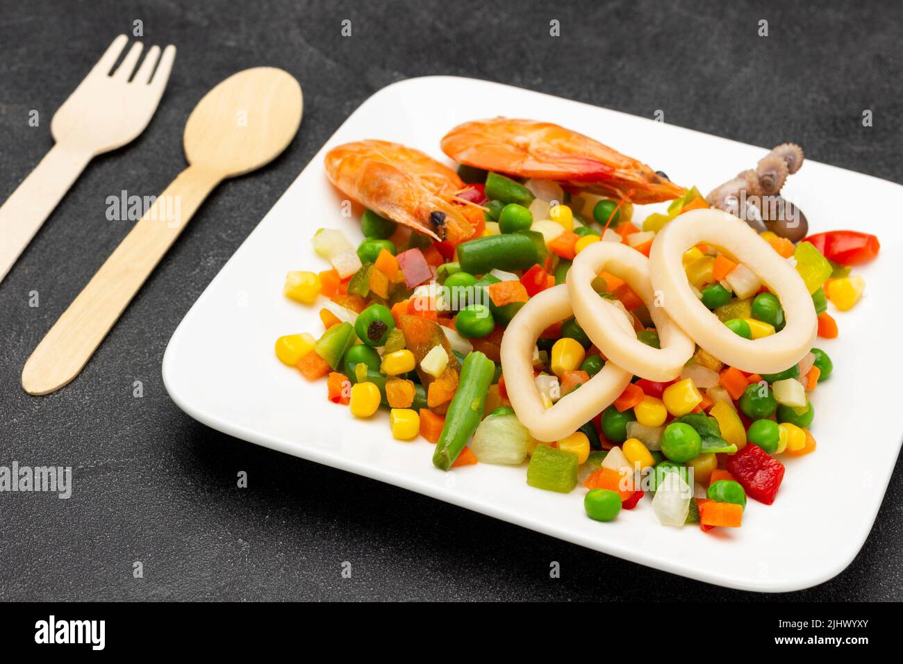 Anillos de camarones y calamares con verduras mezcladas sobre un plato blanco. Cuchara y tenedor sobre la mesa. Vista superior. Fondo negro Foto de stock