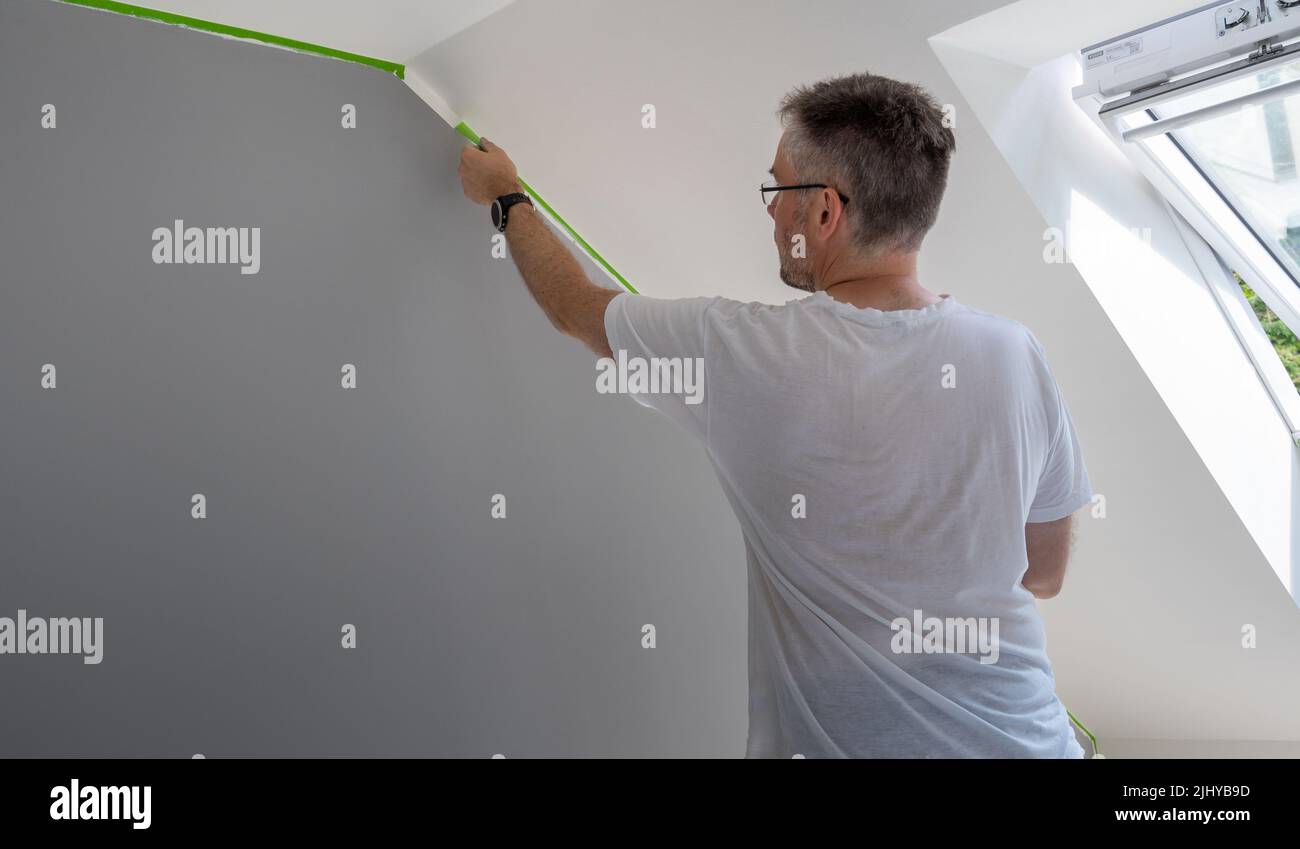 Painter elimina la cinta de enmascarar y crea un borde afilado entre una parte pintada gris y blanca de una pared. Foto de stock