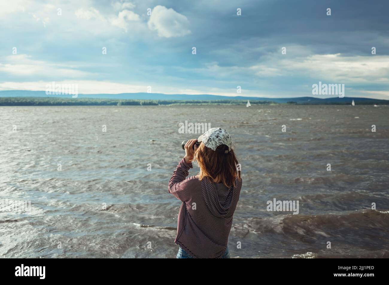 Mujer joven en CAP mirando a través de binoculares a aves en el lago o río Observación de aves, zoología, ecología Investigación en la naturaleza, observación de animales Foto de stock