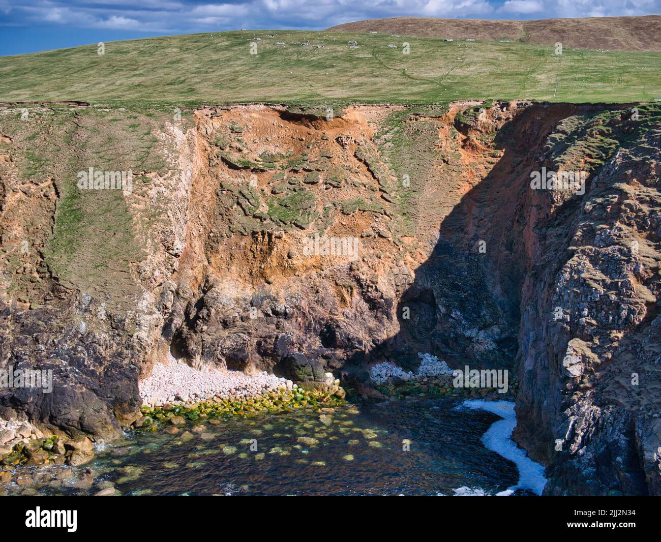 Pérdida de tierras costeras causada por la erosión de la tierra y el deslizamiento hacia el mar. Tomada cerca de Uyea en Northmavine, Shetland, Reino Unido en un día soleado con un cielo azul. Foto de stock