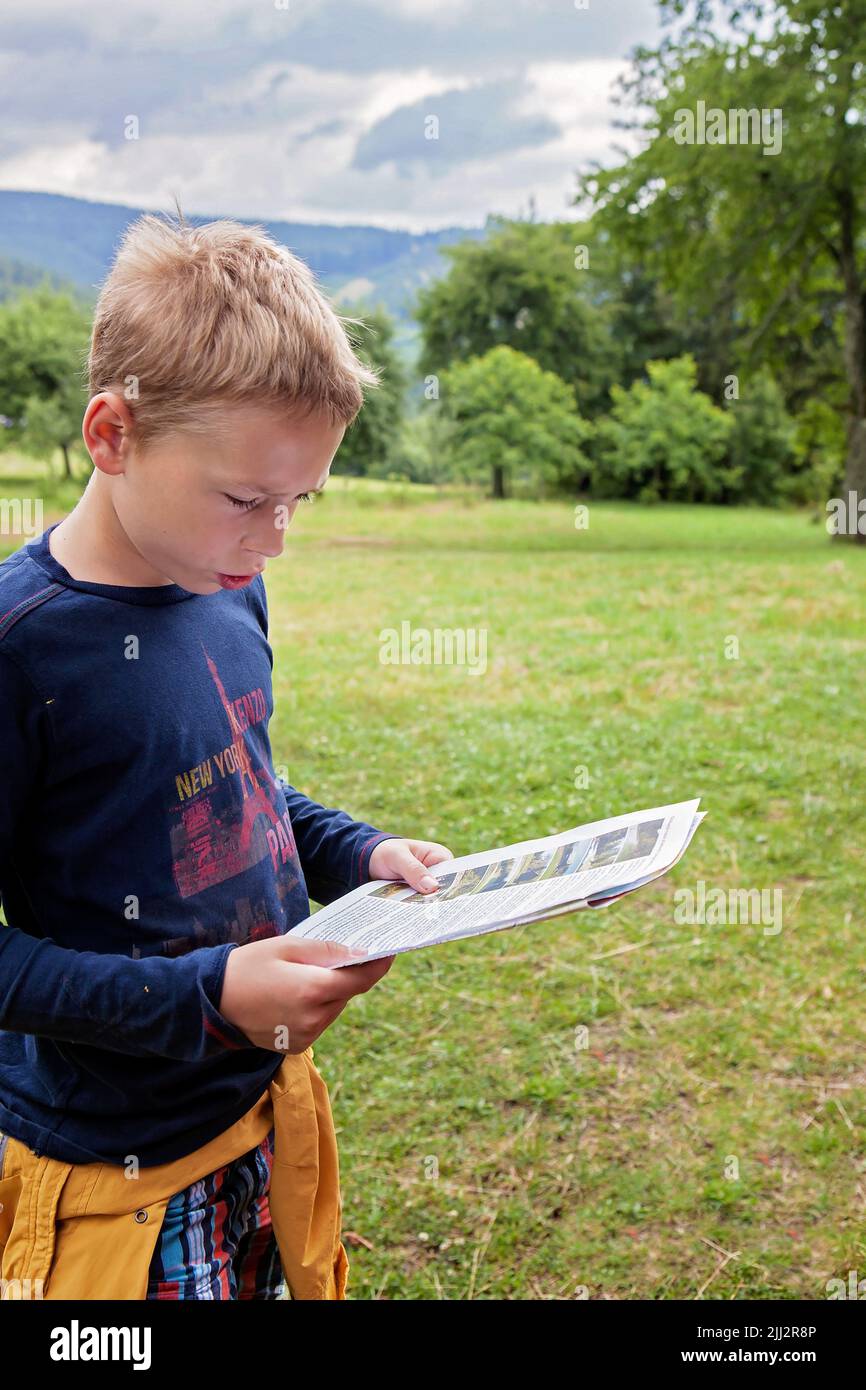 Muchacho joven, adolescente, preadolescente, leyendo un papel, carta con un paisaje de la naturaleza detrás de él. foto natural de un niño rubio caucásico, preguntándose, leyendo, th Foto de stock