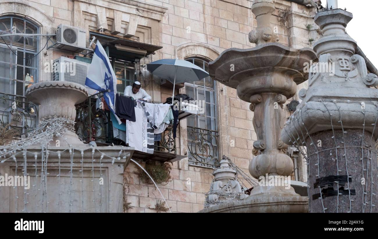Un colono judío cuelga ropa en un balcón adornado con la bandera israelí en el área de Muristán que es un complejo de calles y tiendas en el Barrio Cristiano de la Ciudad Vieja el 20 de julio de 2022 en Jerusalén, Israel. Anteriormente, el Patriarca Ortodoxo Griego de Jerusalén, el Patriarca Teófilo III, dijo al Presidente de los Estados Unidos, Joe Biden, que las organizaciones de derecha israelíes están haciendo esfuerzos para expulsar a los cristianos de la vieja ciudad de Jerusalén, mediante la confiscación de propiedades. Cerca de 300 colonos judíos ya viven en el Barrio Cristiano. Foto de stock