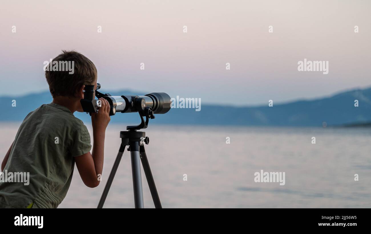 Niño explorando el cielo mirando a través de un telescopio en la noche de pie al lado del mar. Foto de stock
