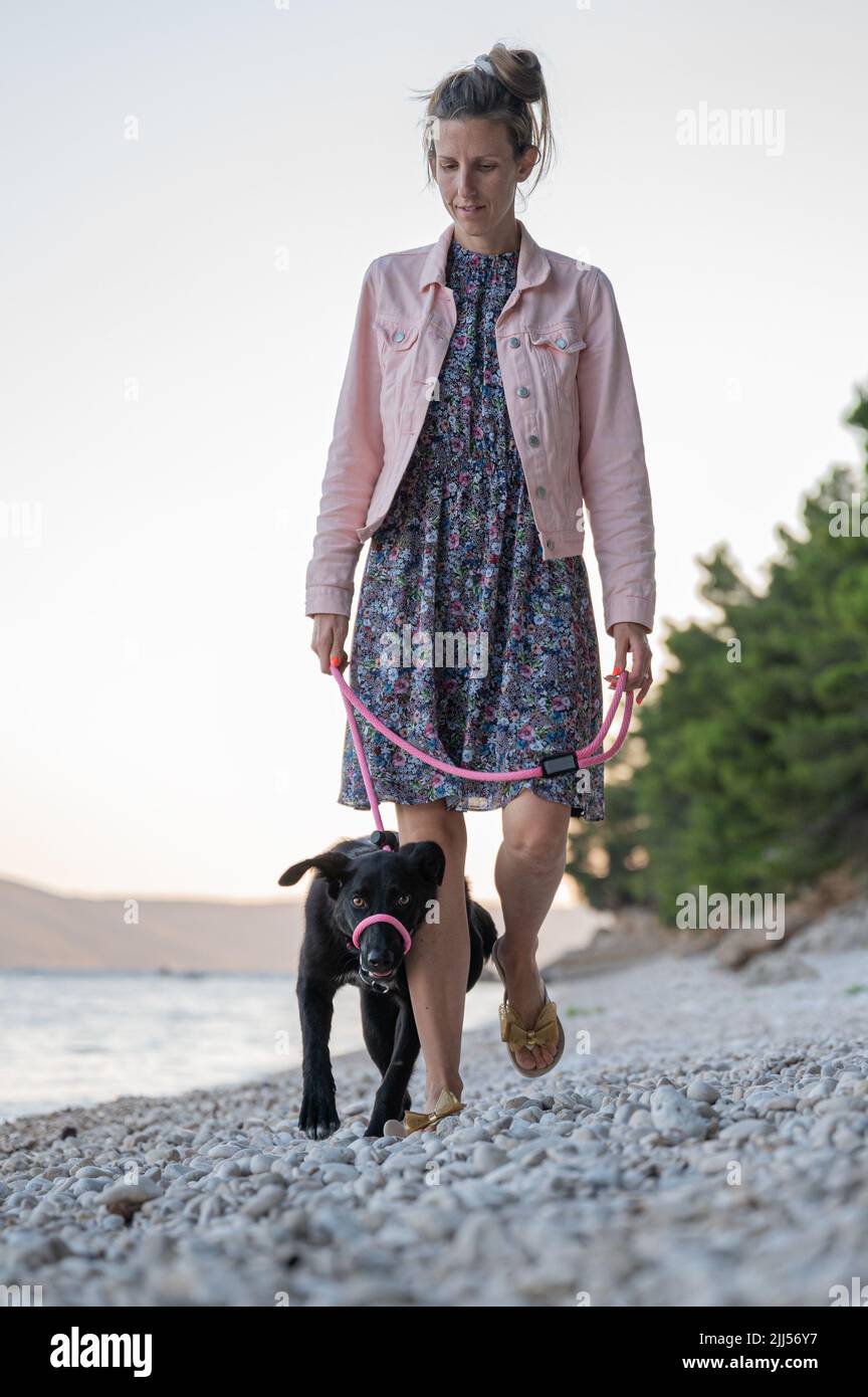 Joven propietaria caminando er perro negro en una correa en una hermosa playa de guijarros. Foto de stock