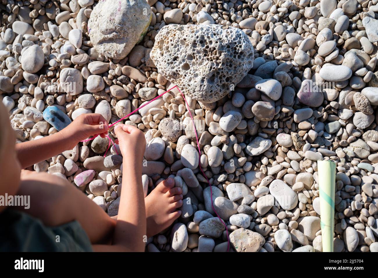 Vista superior de un niño que hace punto una pulsera de cuerda con hilo de algodón de colores en una playa de guijarros. Foto de stock