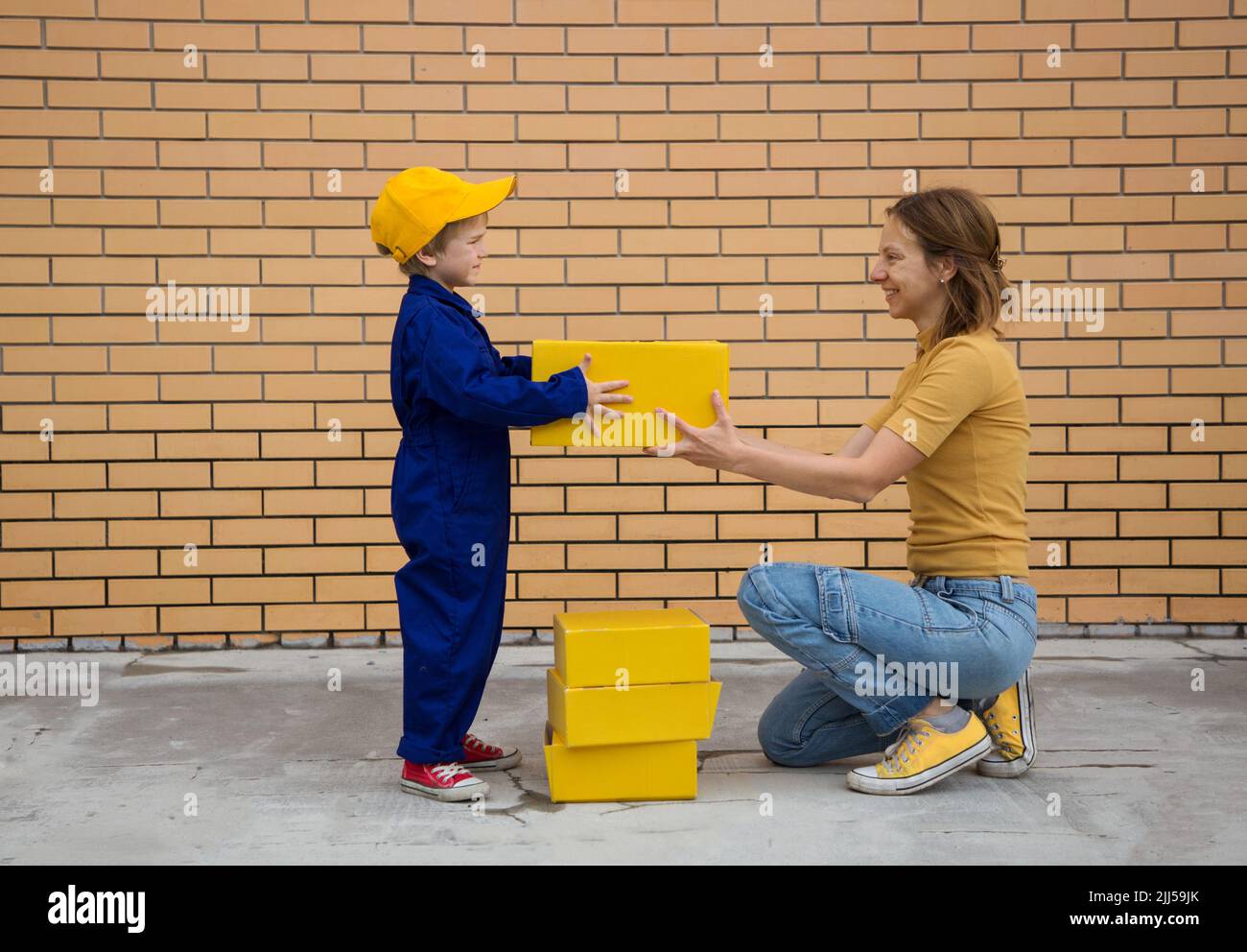 Mujer y niño, madre e hijo pasan las cajas de cartón amarillas de mano en mano. Un pequeño mensajero en el juego del correo. Correo expreso, felicidad de Foto de stock