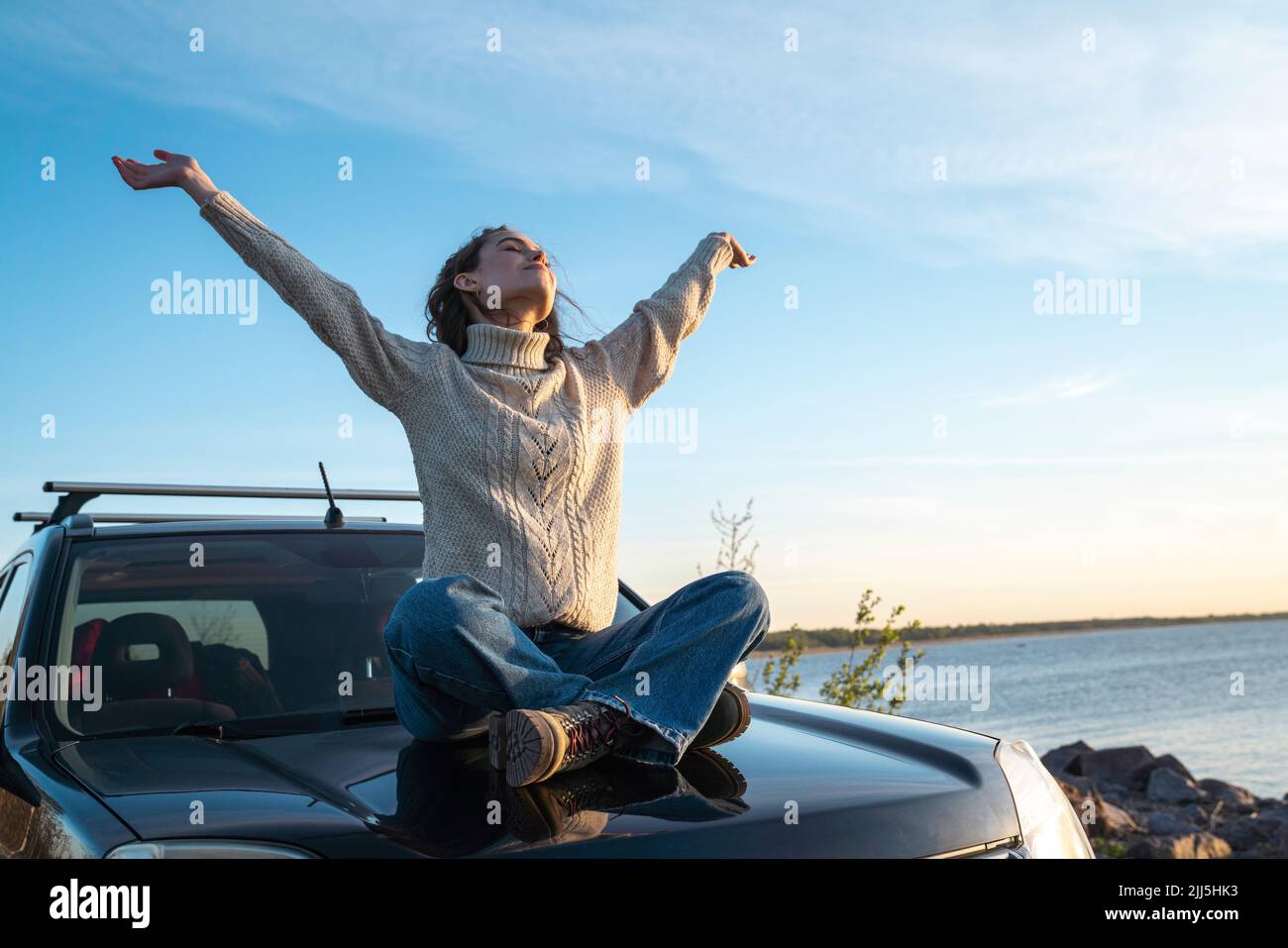 Mujer joven sonriente con los brazos levantados sentada en el capó del coche Foto de stock