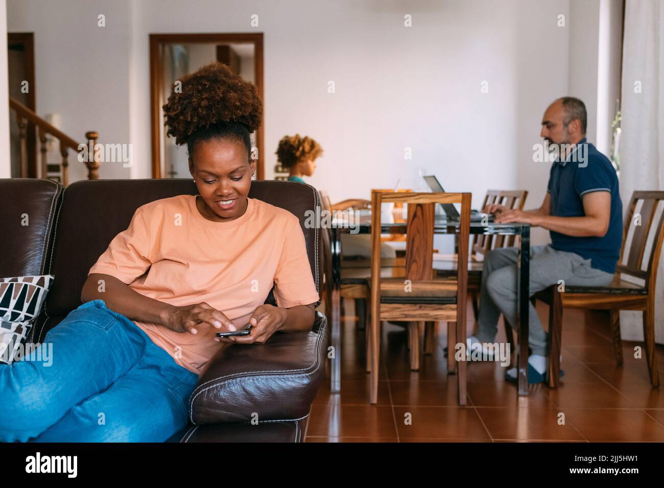 Mujer sonriente con un teléfono inteligente sentada delante de un hombre y una hija en casa Foto de stock