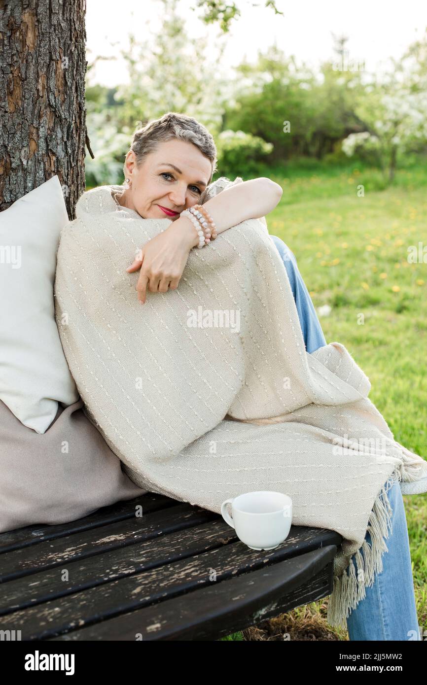 Mujer envuelta en manta sentada con una taza de café en un banco junto al tronco del árbol Foto de stock