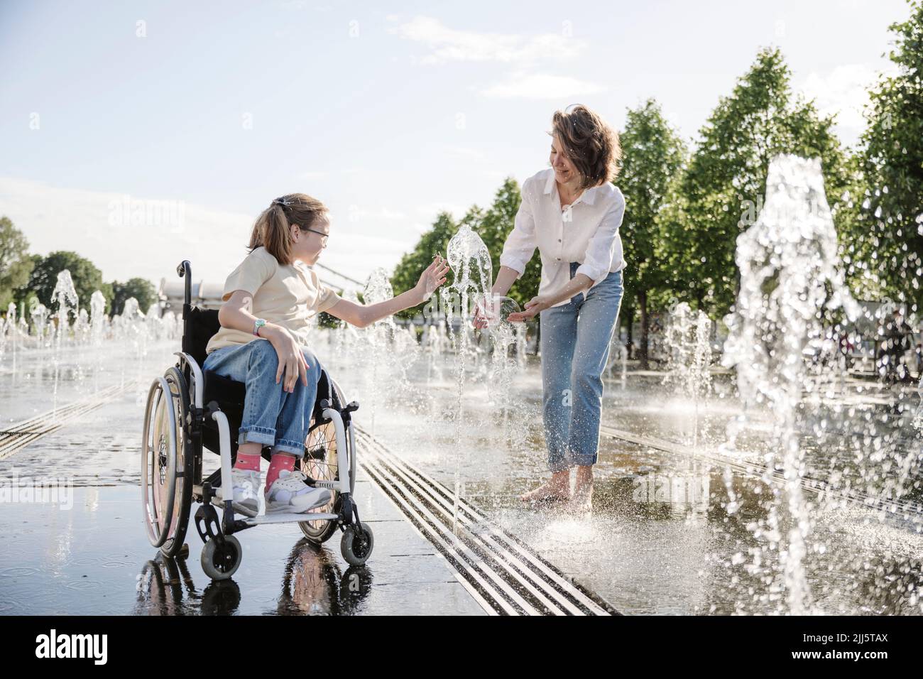 Niña en silla de ruedas jugando junto a la fuente de agua con la madre en el parque Foto de stock
