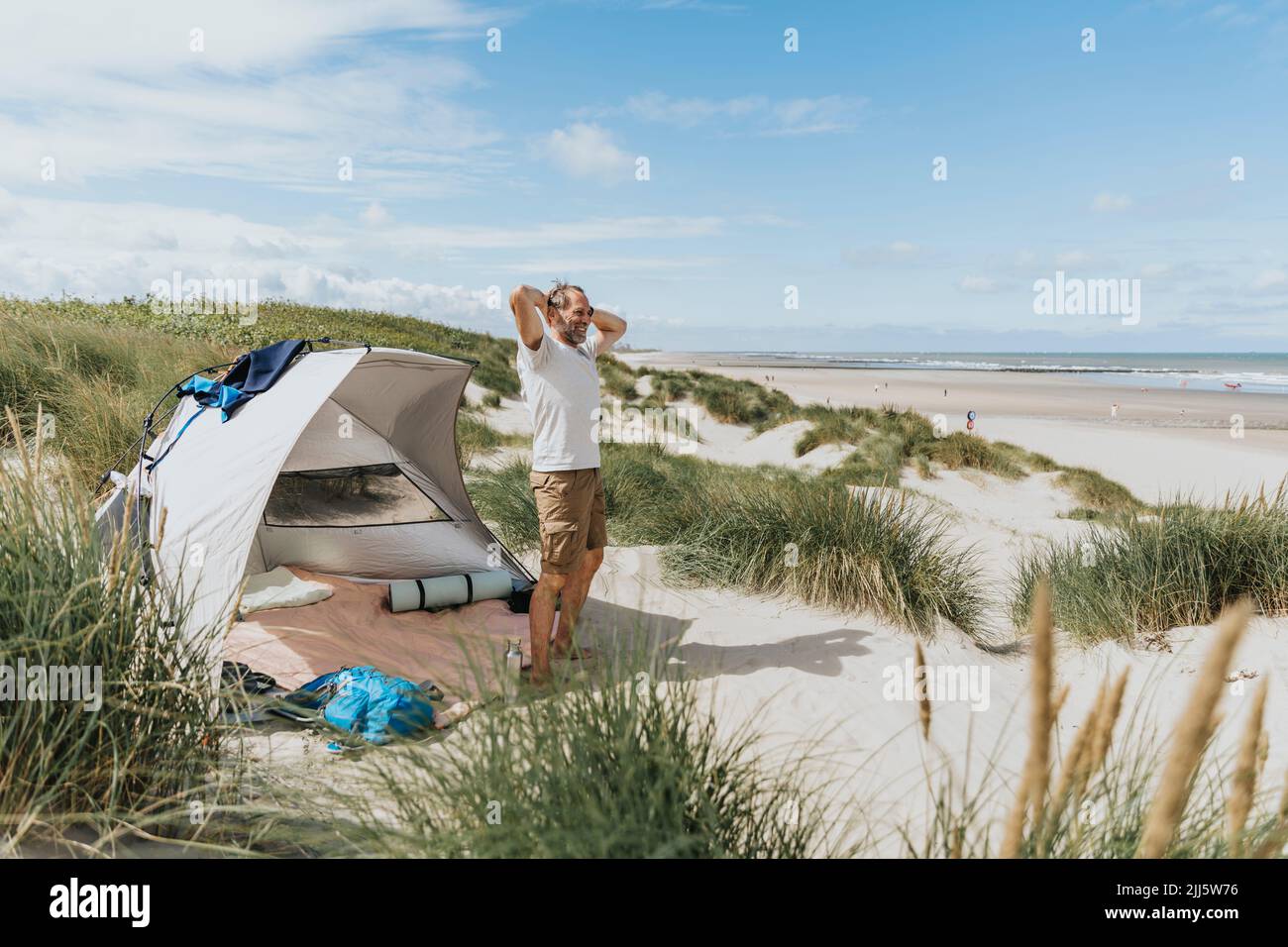 Hombre maduro feliz con las manos detrás de la cabeza de pie en la playa en el día soleado Foto de stock