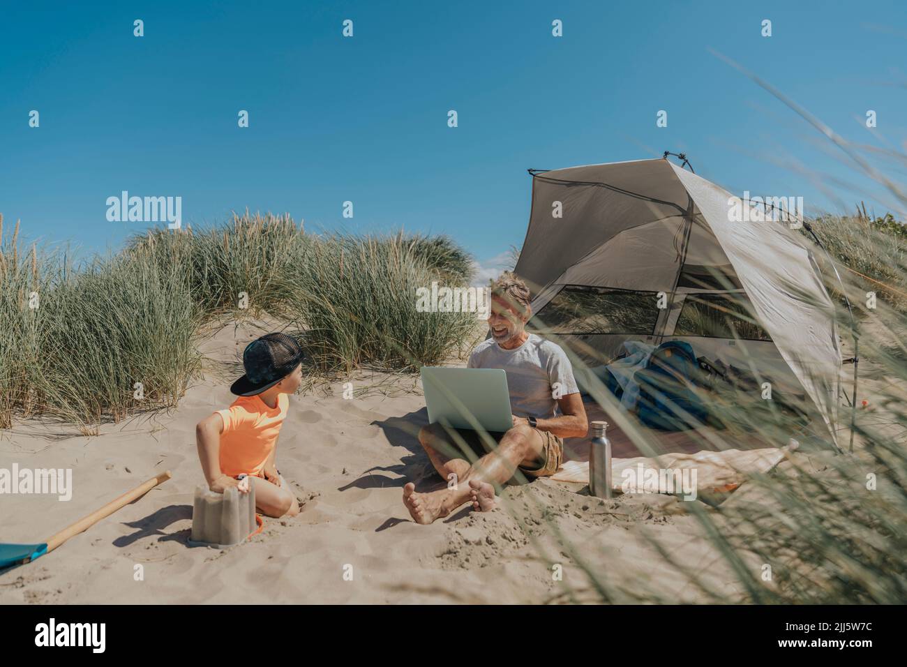 Chico jugando delante de su padre usando el portátil en la playa Foto de stock