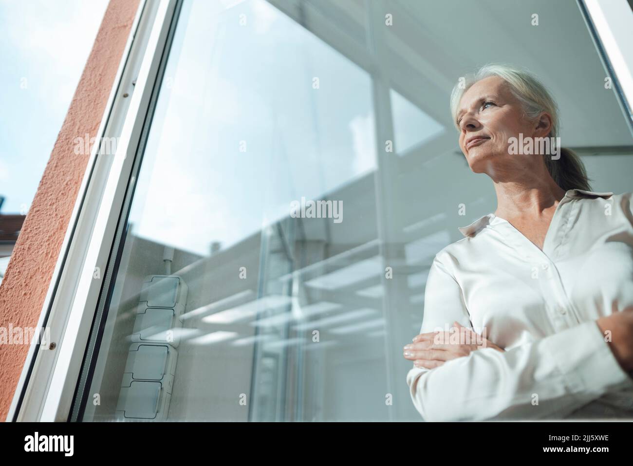 Pensativa mujer de negocios de pie con los brazos cruzados visto a través de vidrio Foto de stock