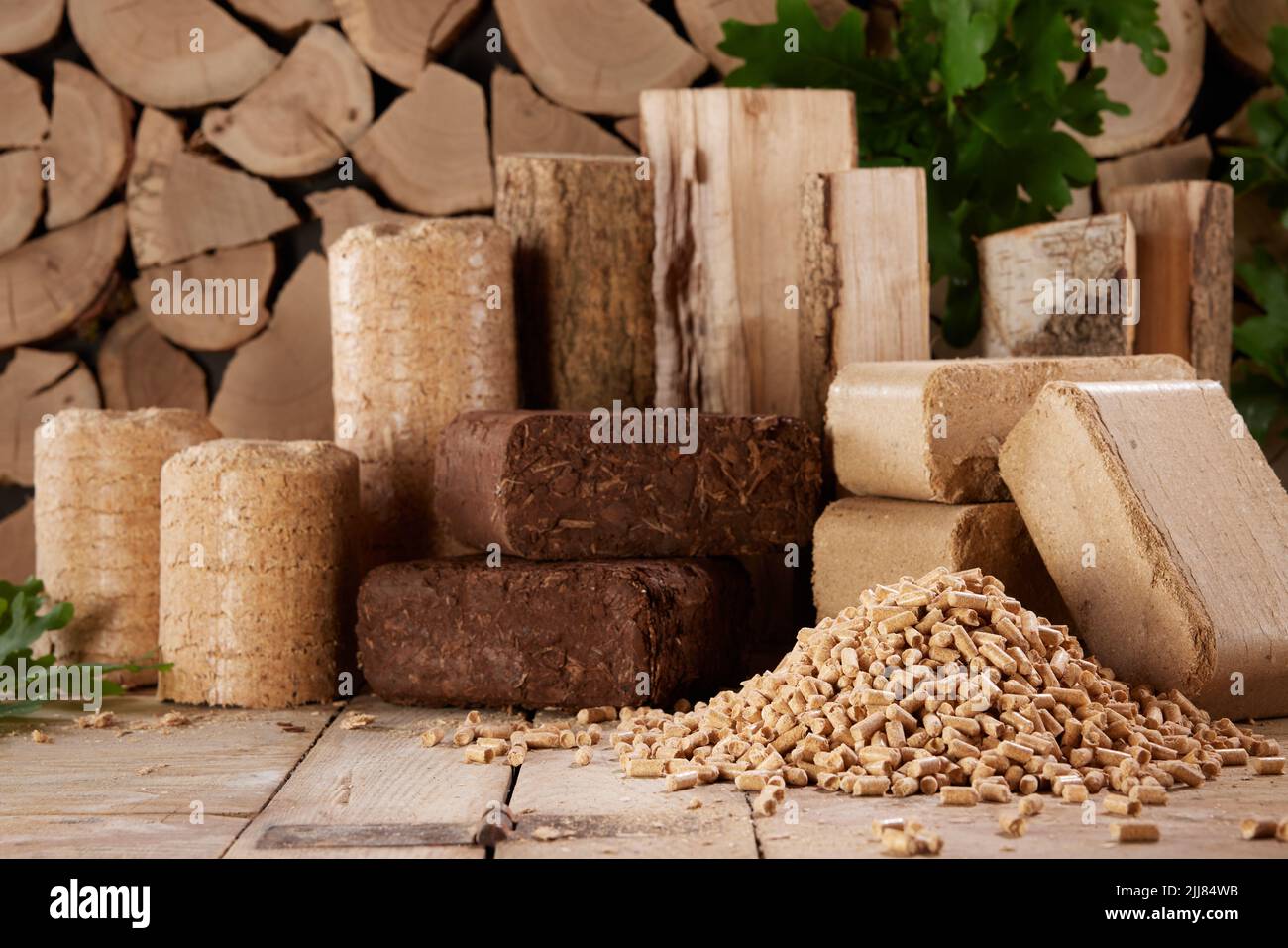 Varias briquetas de biocombustible comprimido y pirámide de pellets de madera dispuestos sobre una mesa de madera contra la pila de leña a la luz del día Foto de stock
