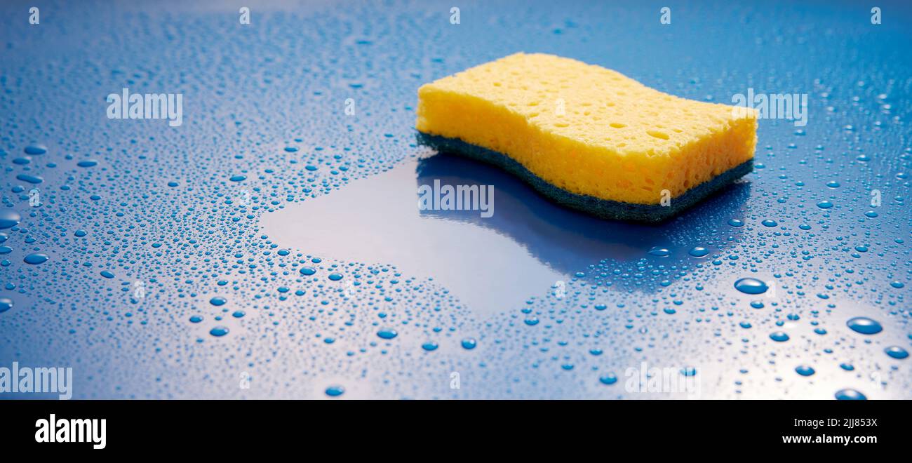 Esponja de color amarillo de limpieza colocada sobre un fondo azul húmedo cubierta con gotas de agua en una sala luminosa durante la rutina de higiene Foto de stock