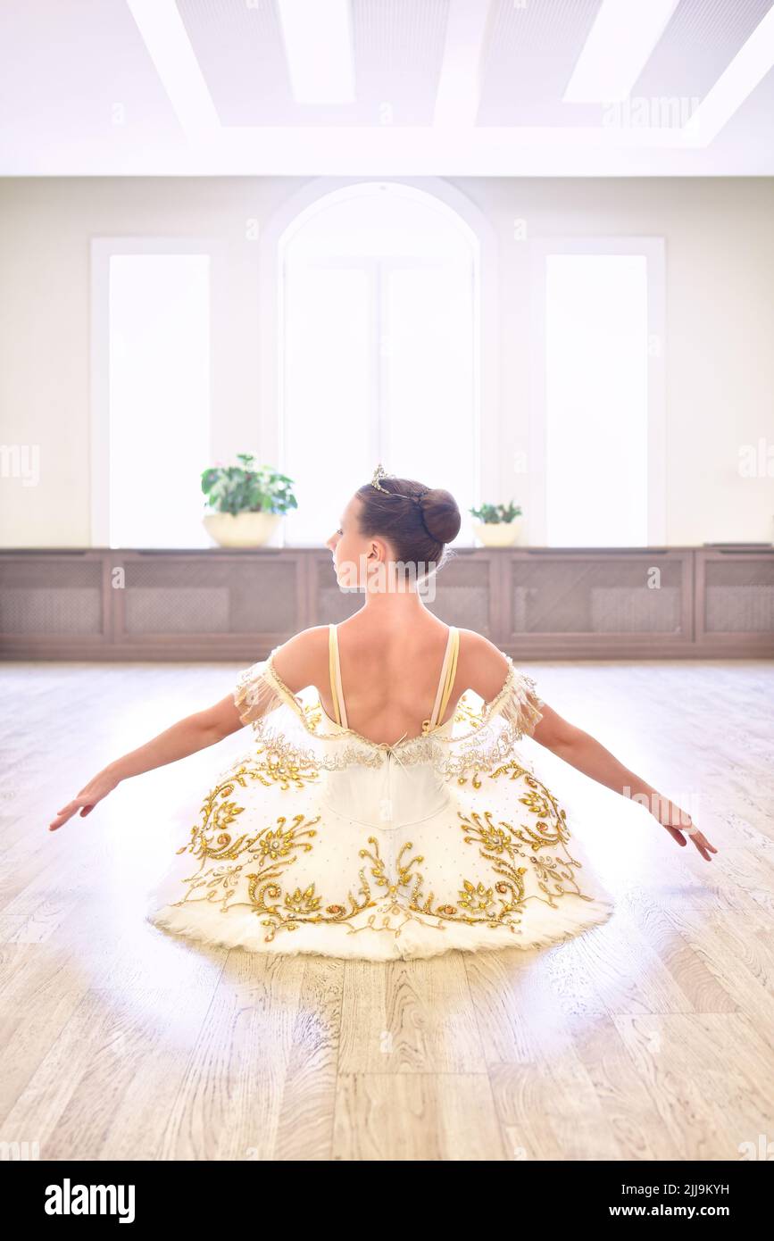 Vista trasera de la joven bailarina de ballet hermosa en vestido de color crema sentado y calienta sus manos en el suelo de madera en el estudio de ballet Foto de stock