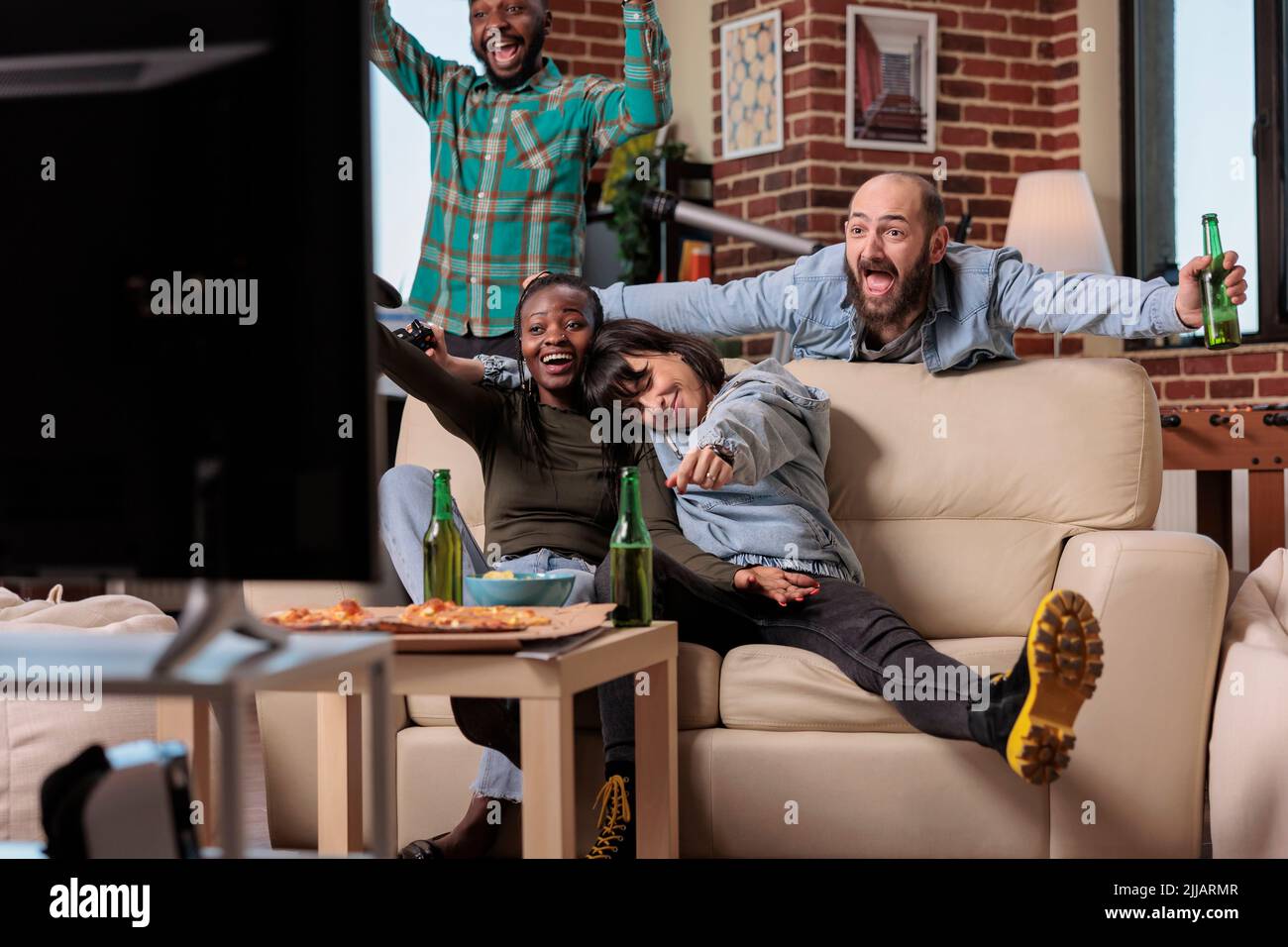 Grupo multiétnico de personas que celebran los videojuegos ganan juntos, jugando a la competencia en una reunión de fiesta con alcohol. La gente se siente feliz por la victoria y ganar, bebiendo cerveza en casa. Foto de stock