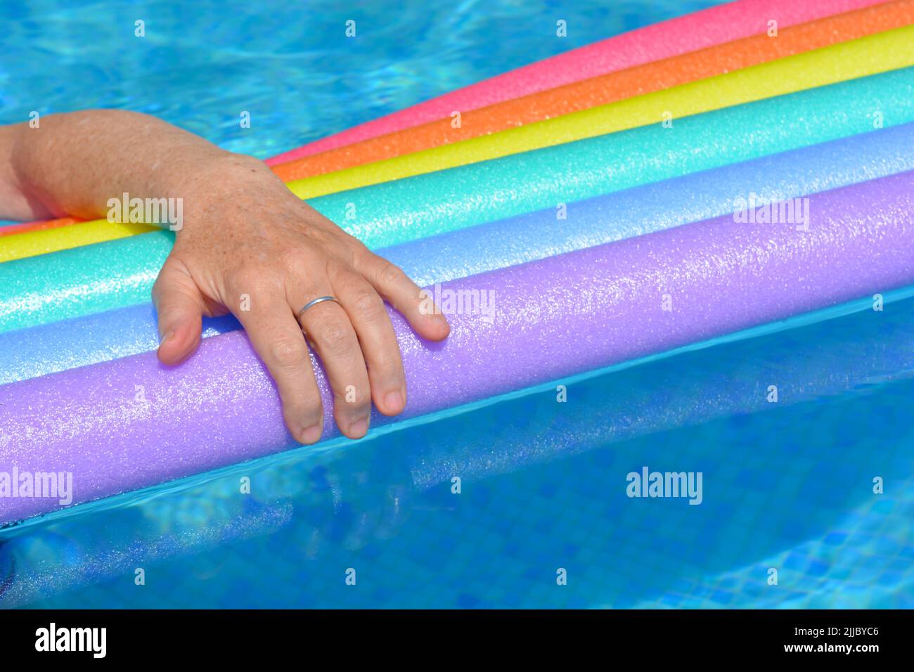 Brazo de mujer descansando sobre fideos de natación de color arco iris flotando en la piscina Foto de stock
