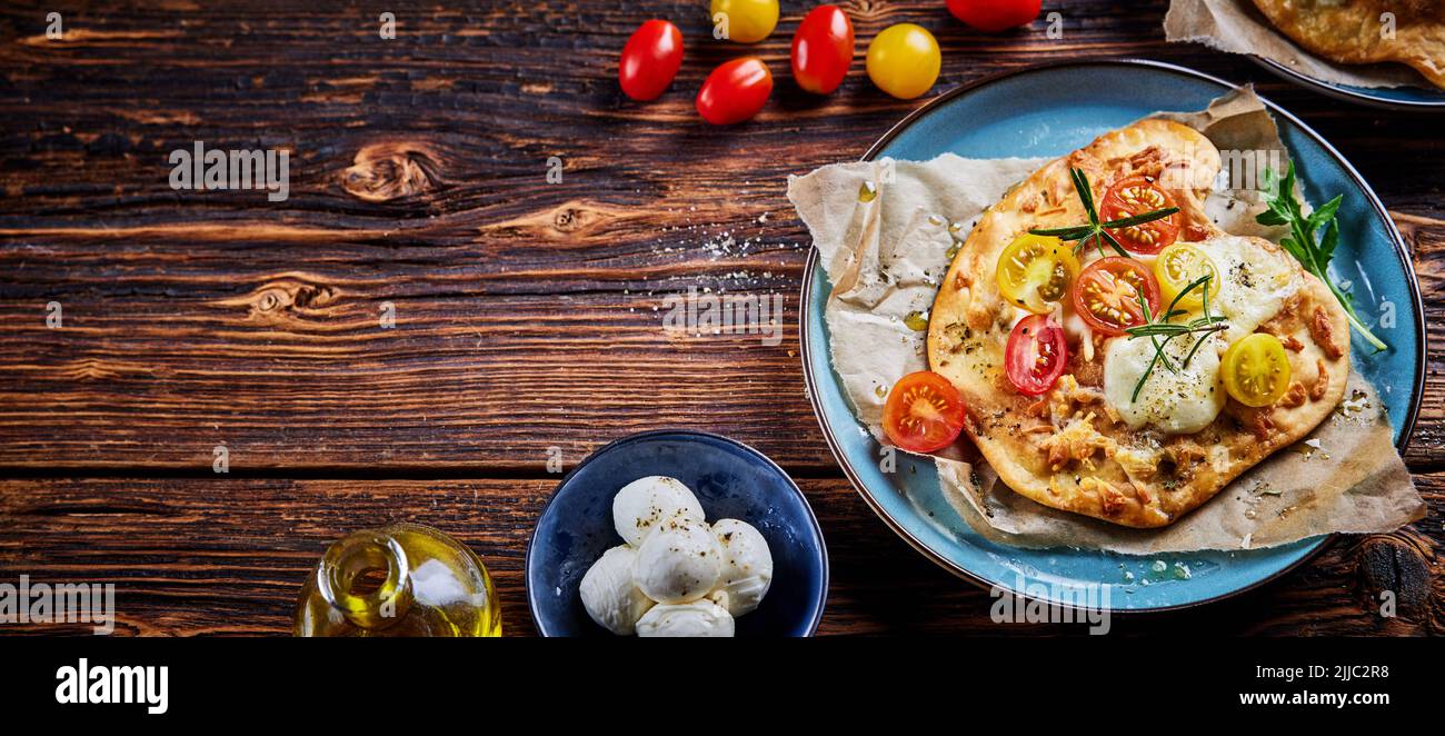Vista superior de deliciosa pizza casera crujiente de pan plano con mozzarella de tomates cherry y hierbas servidas en el plato junto a los ingredientes sobre una mesa de madera Foto de stock