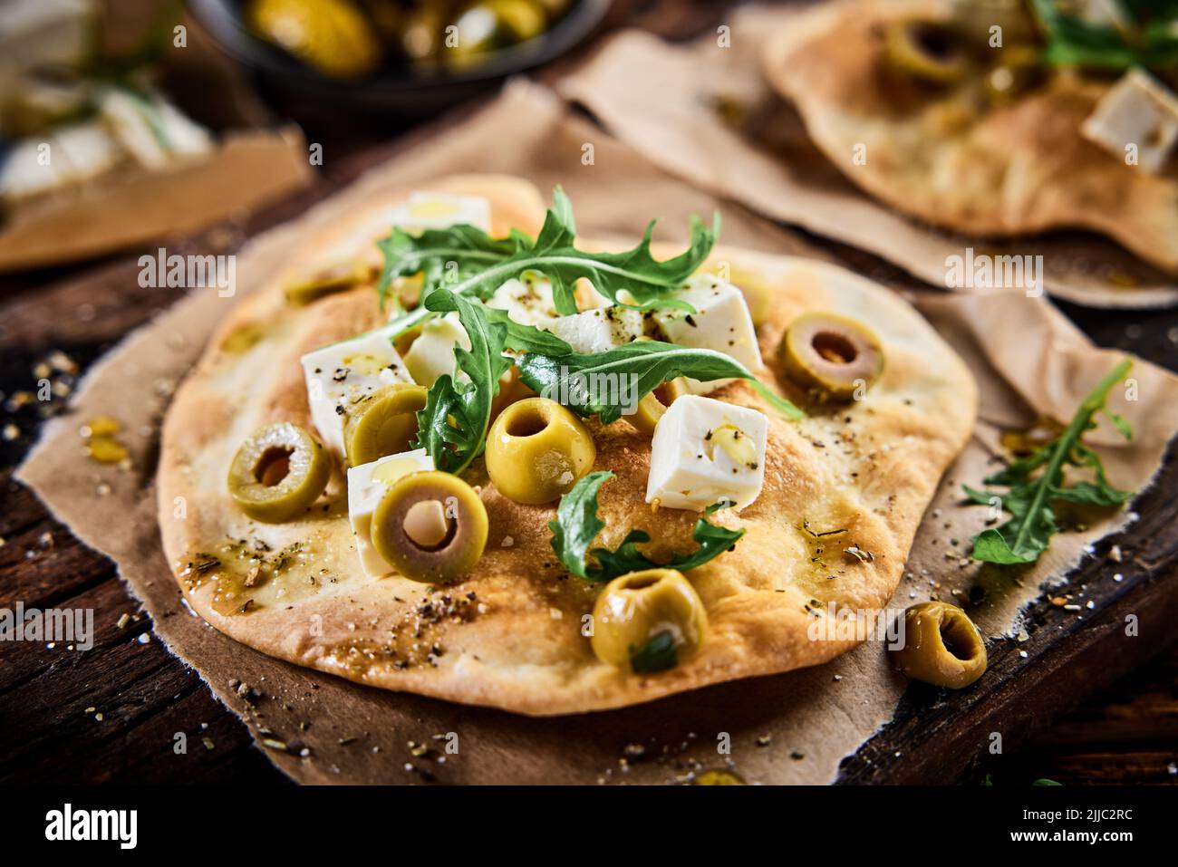Desde arriba de queso feta cortado con aceitunas y rúcula en crujiente pan de pizza servido sobre una tabla de madera en la cocina del restaurante Foto de stock